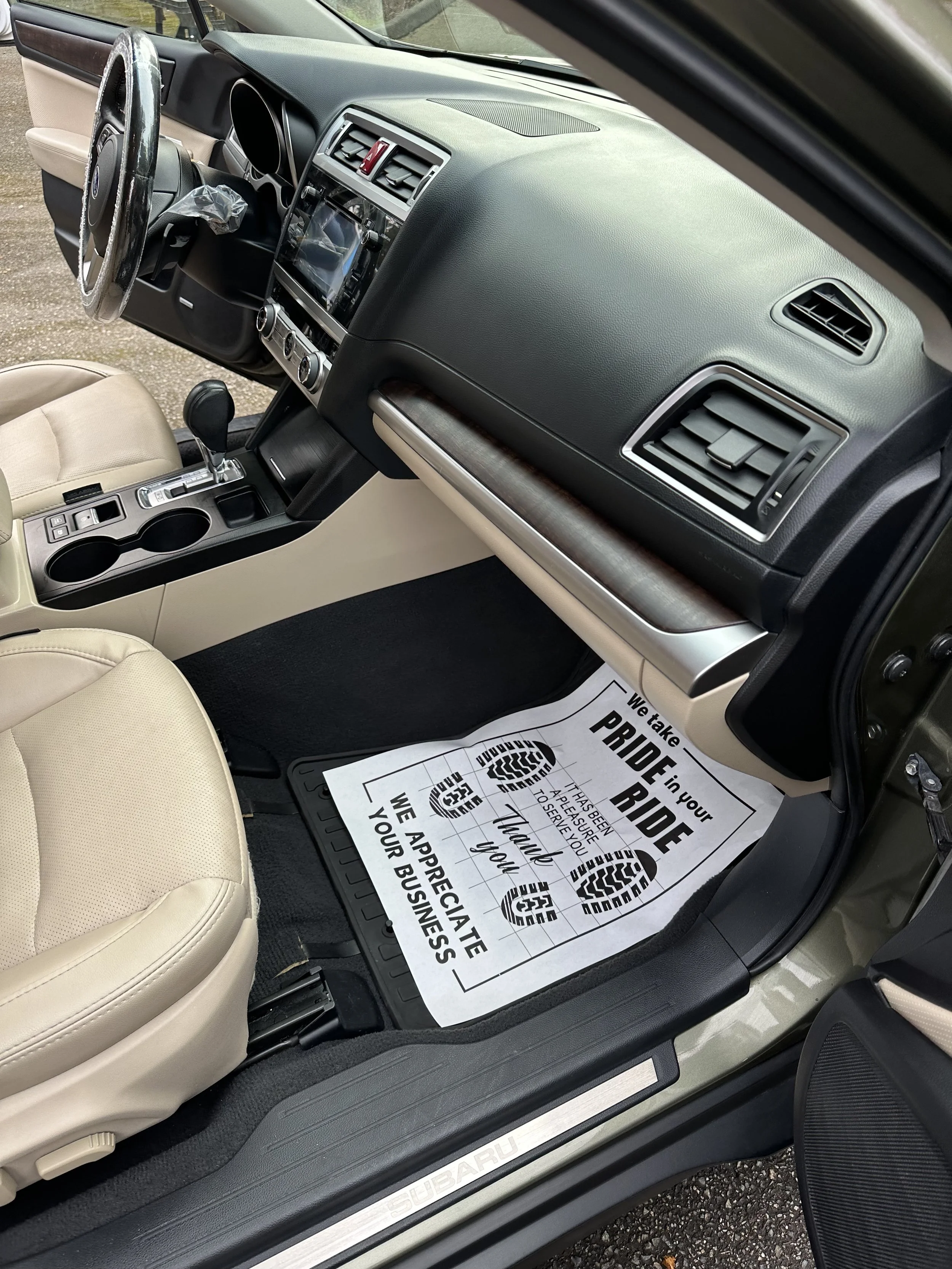 The interior of a Subaru vehicle showing the dashboard, steering wheel, center console with gear shift, cup holders, and a paper floor mat with congratulatory message. The car has beige seats and black interior accents.