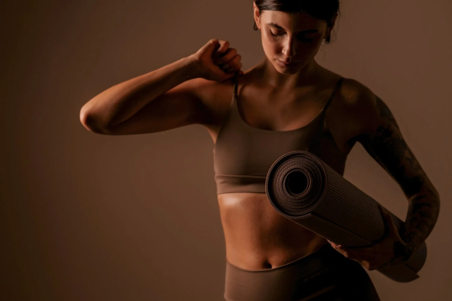 A woman dressed in workout attire holding a yoga mat with a focused expression, in a dimly lit room.