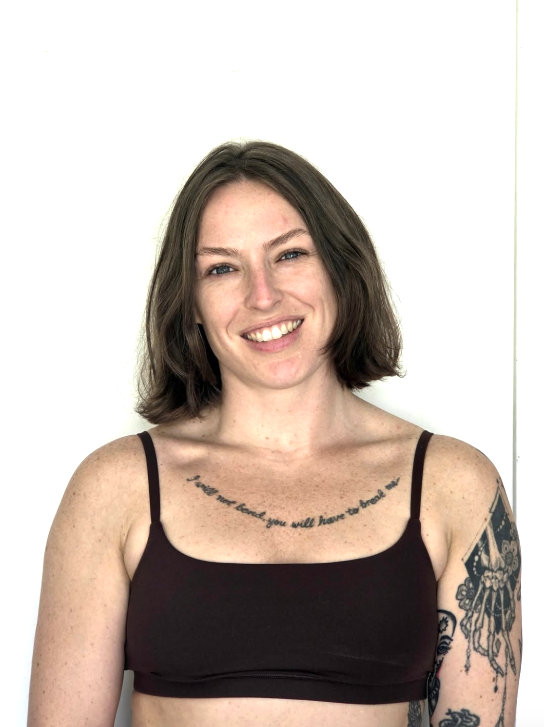 A smiling woman with shoulder-length brown hair, wearing a black spaghetti strap top, standing against a plain white background. She has visible tattoos on her chest and arm.