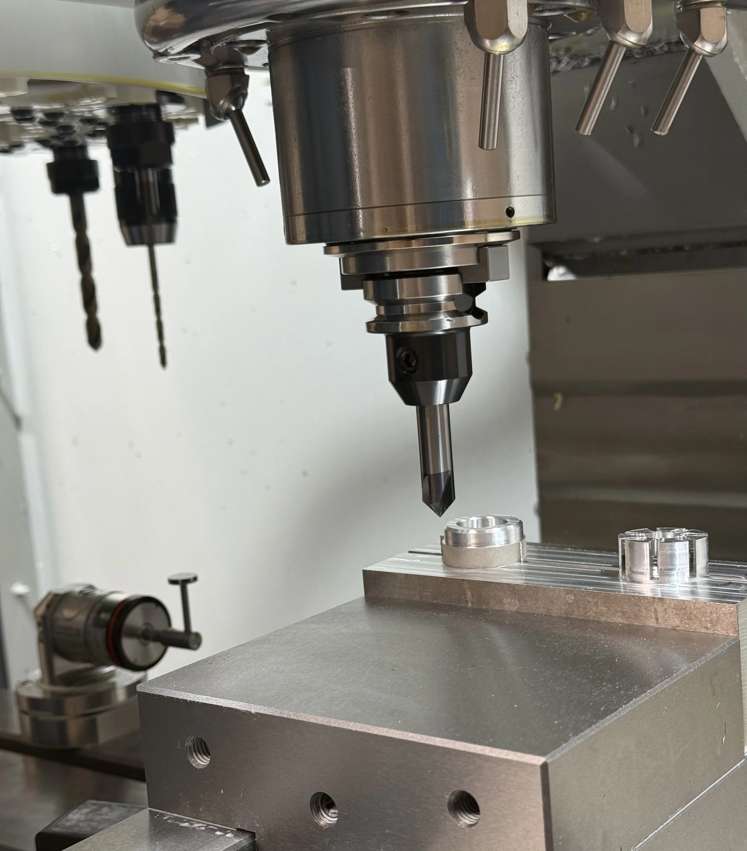 A close-up view of a CNC machine or milling machine with metal drill bits, cutting tools, and a workpiece. The machine's components and work surface are visible, indicating an industrial or manufacturing setting.