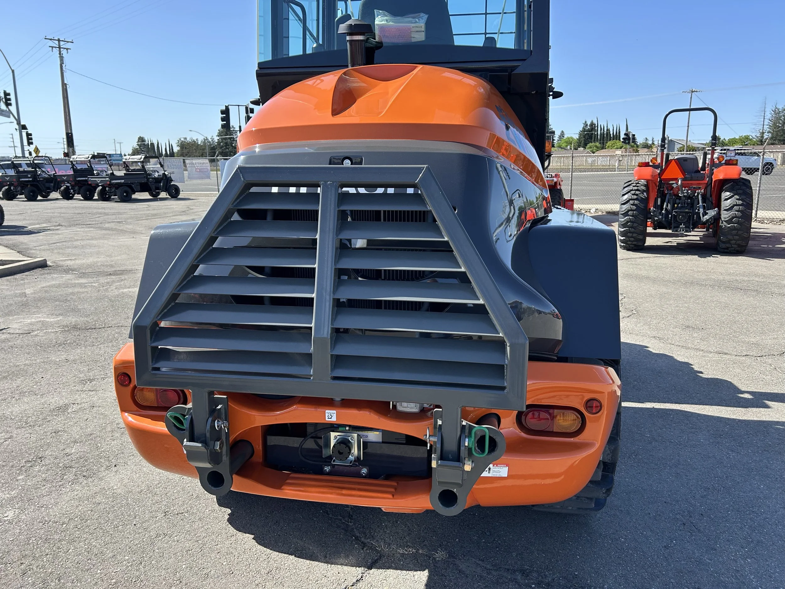 Front view of an orange construction vehicle with a black cab and grill, parked on a concrete lot with utility poles and traffic lights in the background.