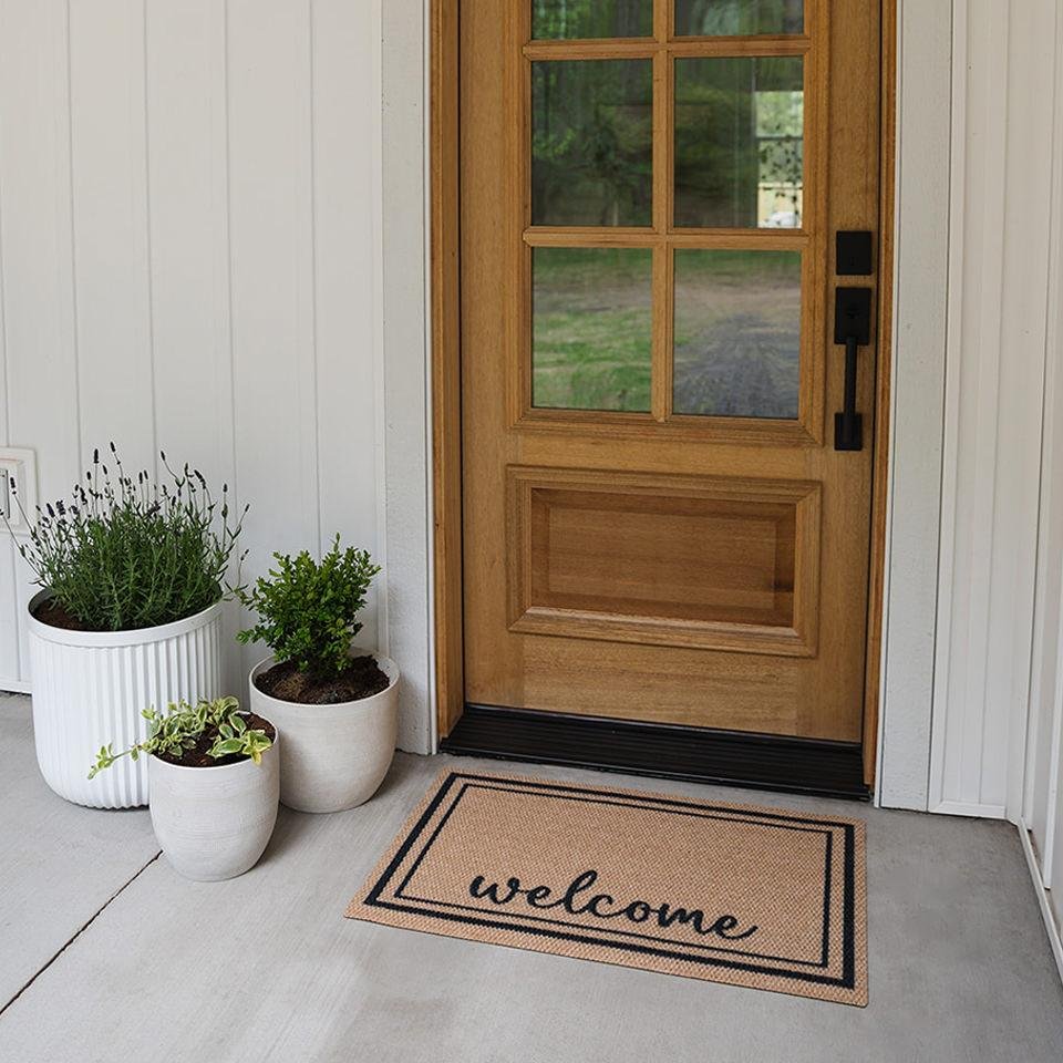 Front porch entrance with a wooden door featuring glass panels and black hardware, a beige welcome mat with black borders and "welcome" written on it, and three potted plants in white and beige containers to the left of the door, set on a concrete floor.