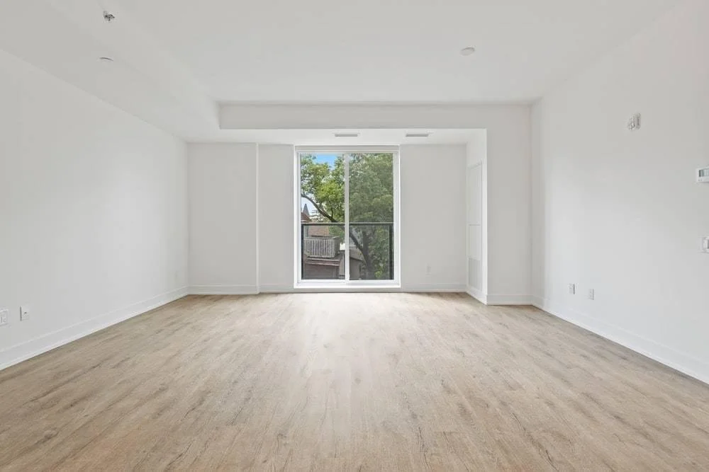 Empty room with white walls, light wood flooring, and a large window showing trees outside.