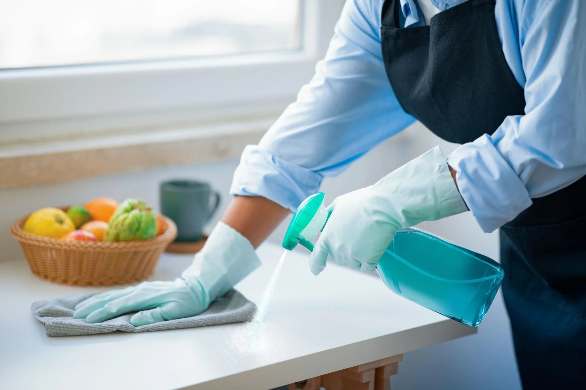 Person wearing gloves cleaning a kitchen counter with disinfectant spray, with a basket of fruit in the background.