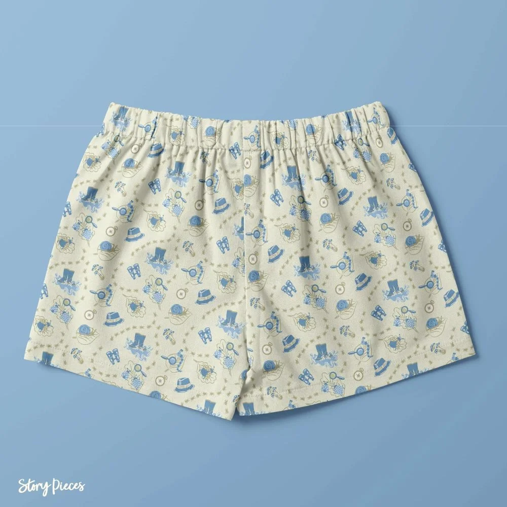 Children’s shorts featuring a playful illustrated pattern of snails, insects, magnifying glasses, boots, and hats in soft blue tones on a light background, set against a blue backdrop.