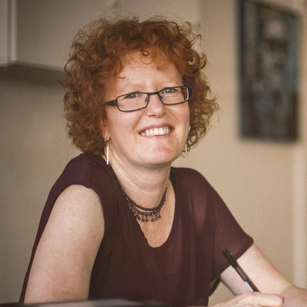 Portrait of Sue Cheer, surface pattern designer, smiling at a desk with a pen in hand, natural light and relaxed studio setting.