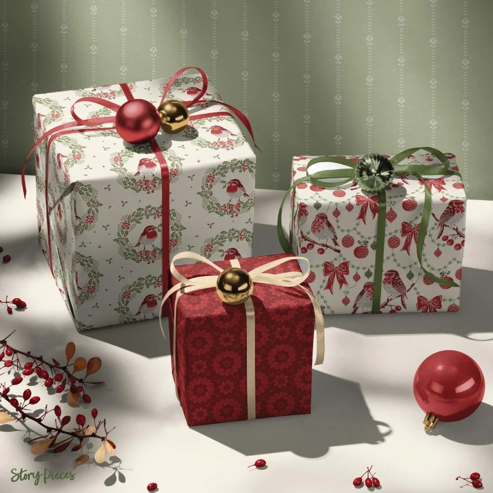 Three wrapped Christmas presents displayed on a table, featuring festive patterned paper with birds, wreaths, and ornaments, tied with red, green, and cream ribbons, surrounded by baubles and winter foliage against a soft green background.