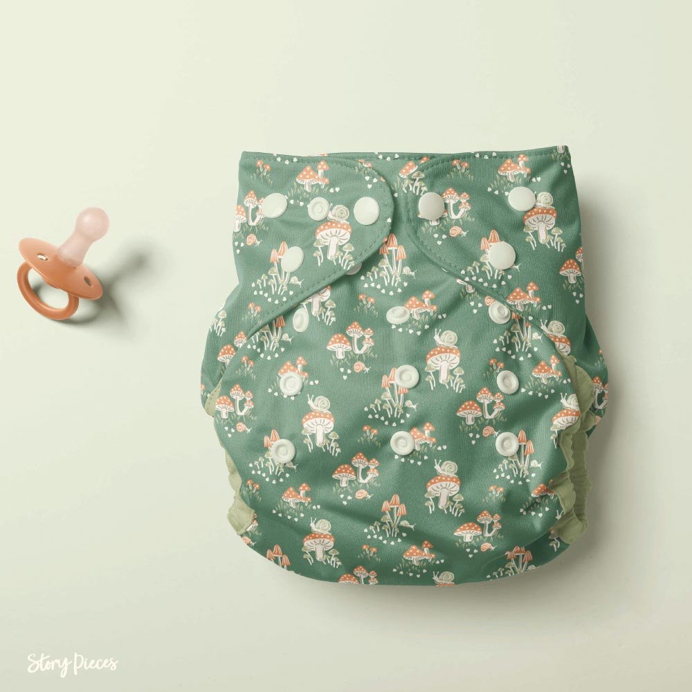 Reusable cloth diaper featuring a whimsical woodland mushroom and snail pattern on a soft green background, shown with snap fastenings alongside a peach-coloured pacifier.