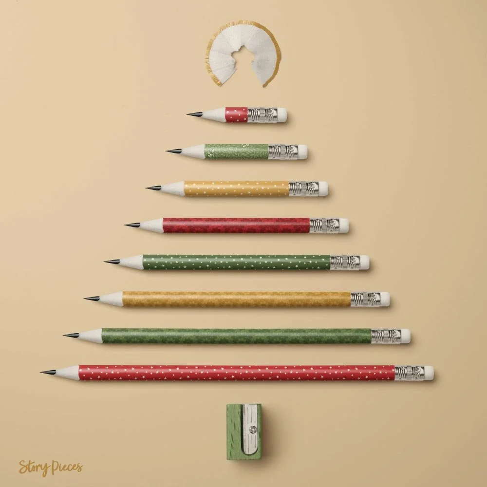 A flat lay of eight pencils arranged by size on a warm neutral background, each decorated with different festive patterns in red, green, and gold from the collection. A curled wood shaving sits above the pencils, with a small green sharpener below, c