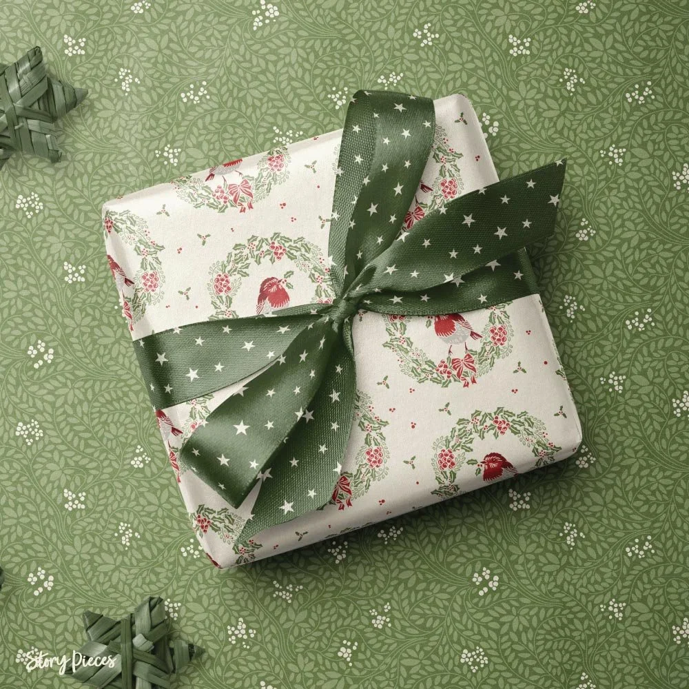 Wrapped Christmas gift in bird-and-wreath patterned paper, tied with a wide green ribbon with white stars, placed on a leafy green patterned background.