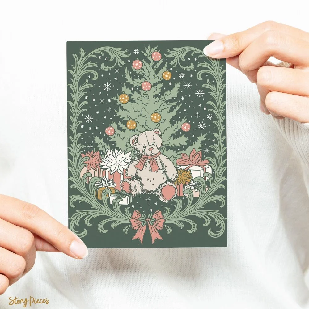 A person holds a square Christmas card featuring a hand-drawn illustration of a teddy bear sitting beneath a decorated tree, surrounded by wrapped gifts and framed by ornate foliage, on a deep green background with soft festive colours.