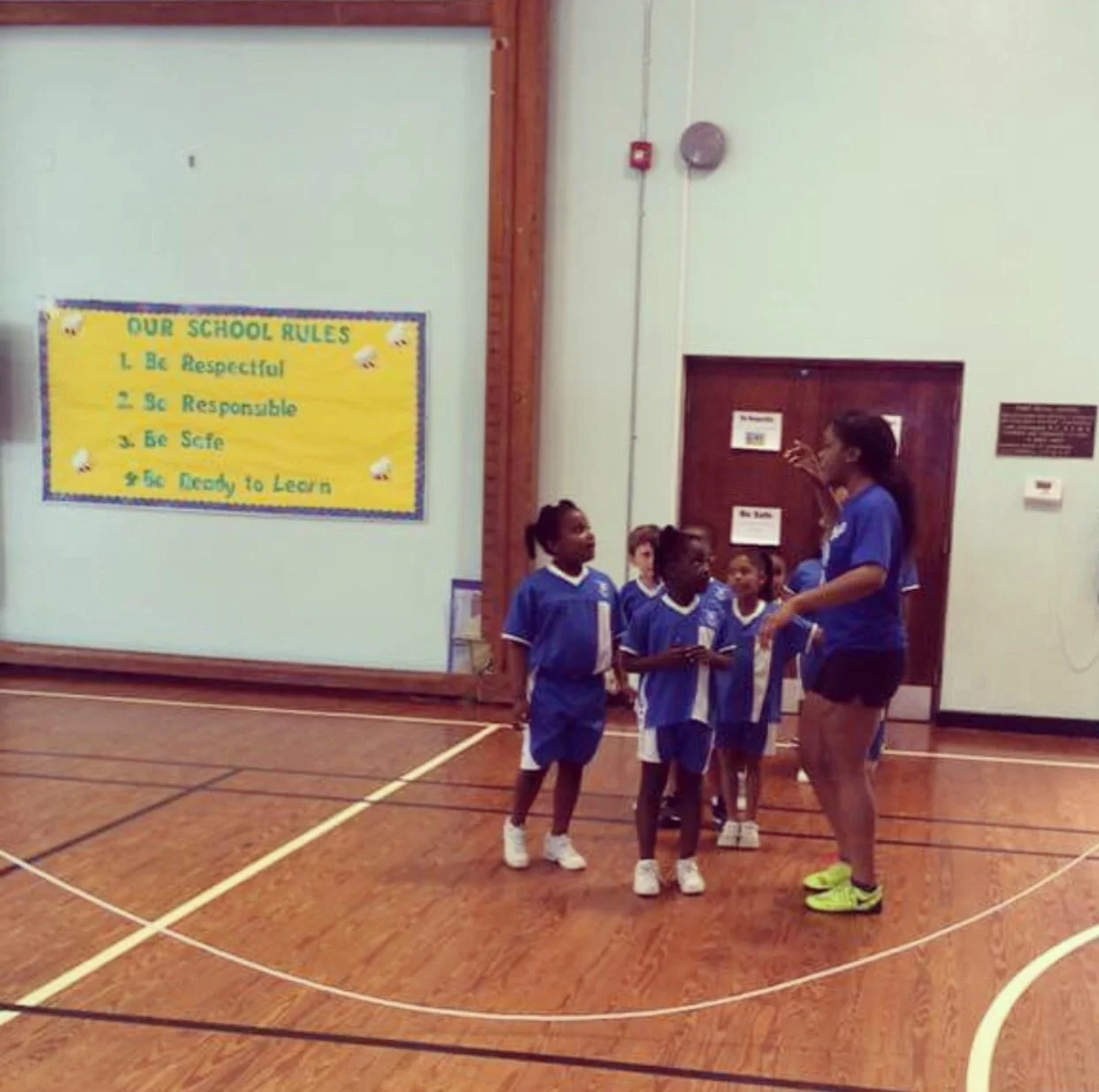 A group of young girls in blue soccer uniforms listening to a woman in a blue shirt and black shorts inside a gymnasium. A yellow bulletin board with school rules is visible on the wall behind them.