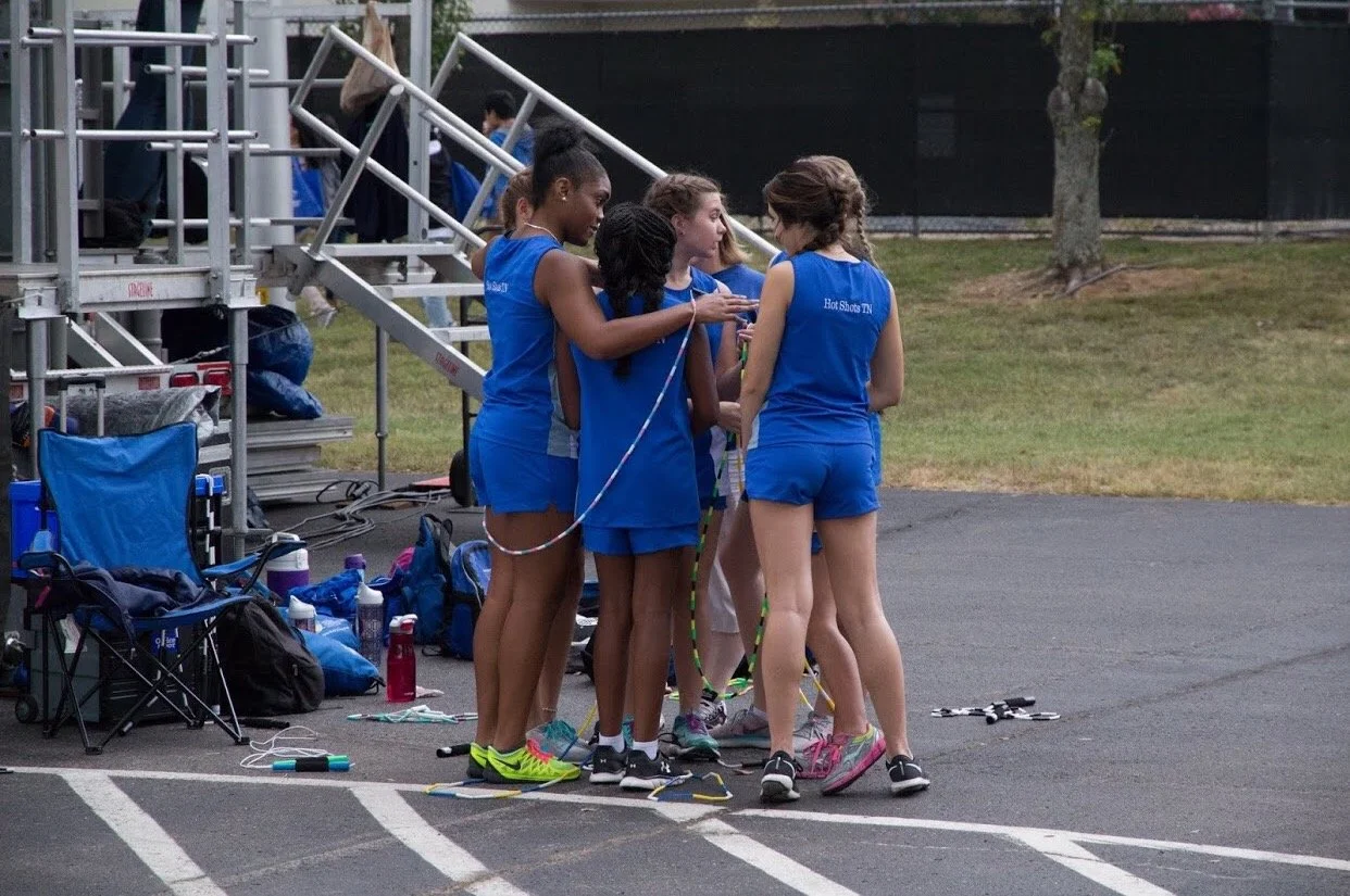Group of young female athletes in blue uniforms huddled together on a track field, preparing for a relay race.