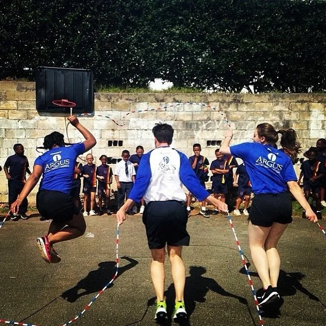 Children playing jump rope outdoors, with two girls jumping simultaneously and a boy turning the rope.