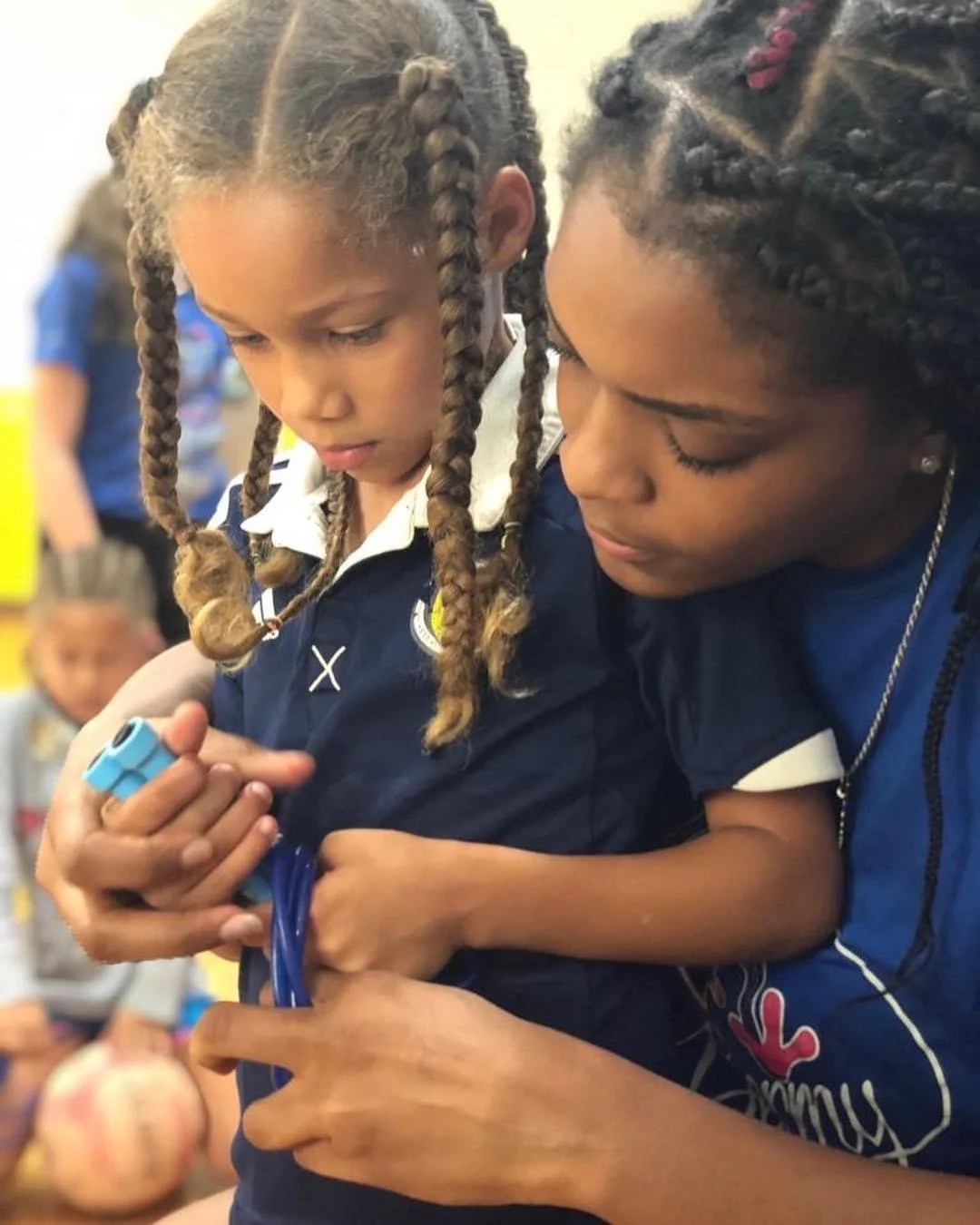 Two young girls with braided hair examining a small electronic device together in a classroom or school setting.