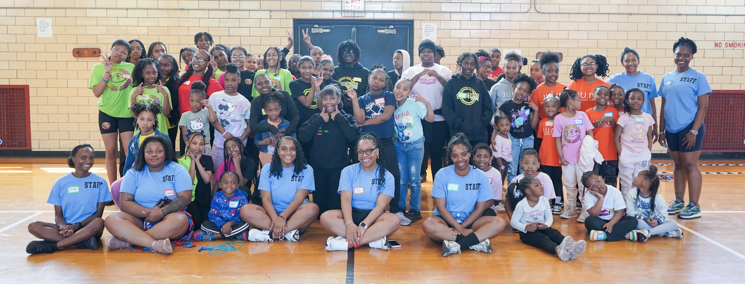 Group of children and adults posing for a photo in a gymnasium with brick walls and a wooden floor, some children sitting on the floor and some standing, with a few adults wearing blue staff shirts.