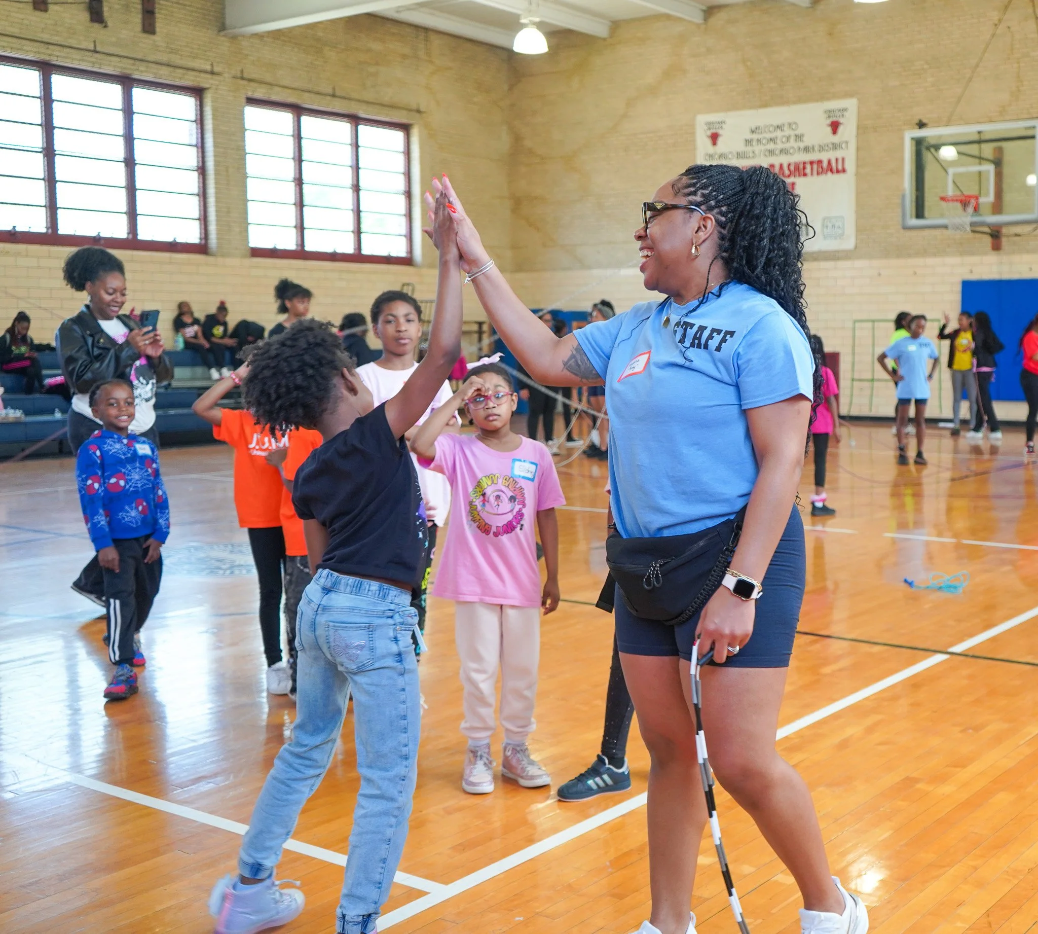 Coach Treyanna with braided hair and glasses giving a high five to a young girl at an Illinois Jump Rope event in a gymnasium filled with children and adults.