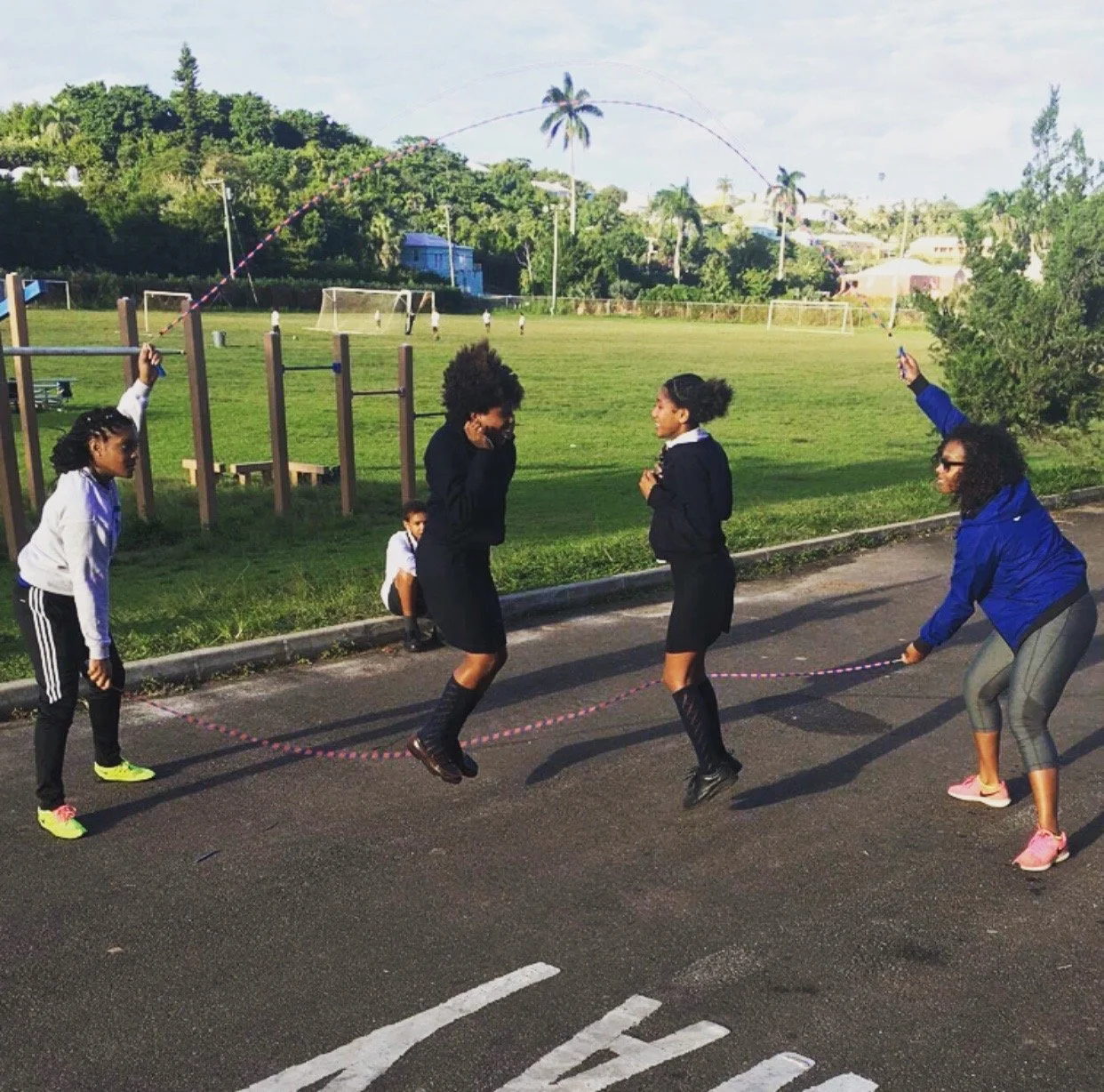 Four women and a girl skipping rope in a parking lot near a grass field with playground equipment and a soccer goal in the background. Two women are jumping, and two are holding the rope.