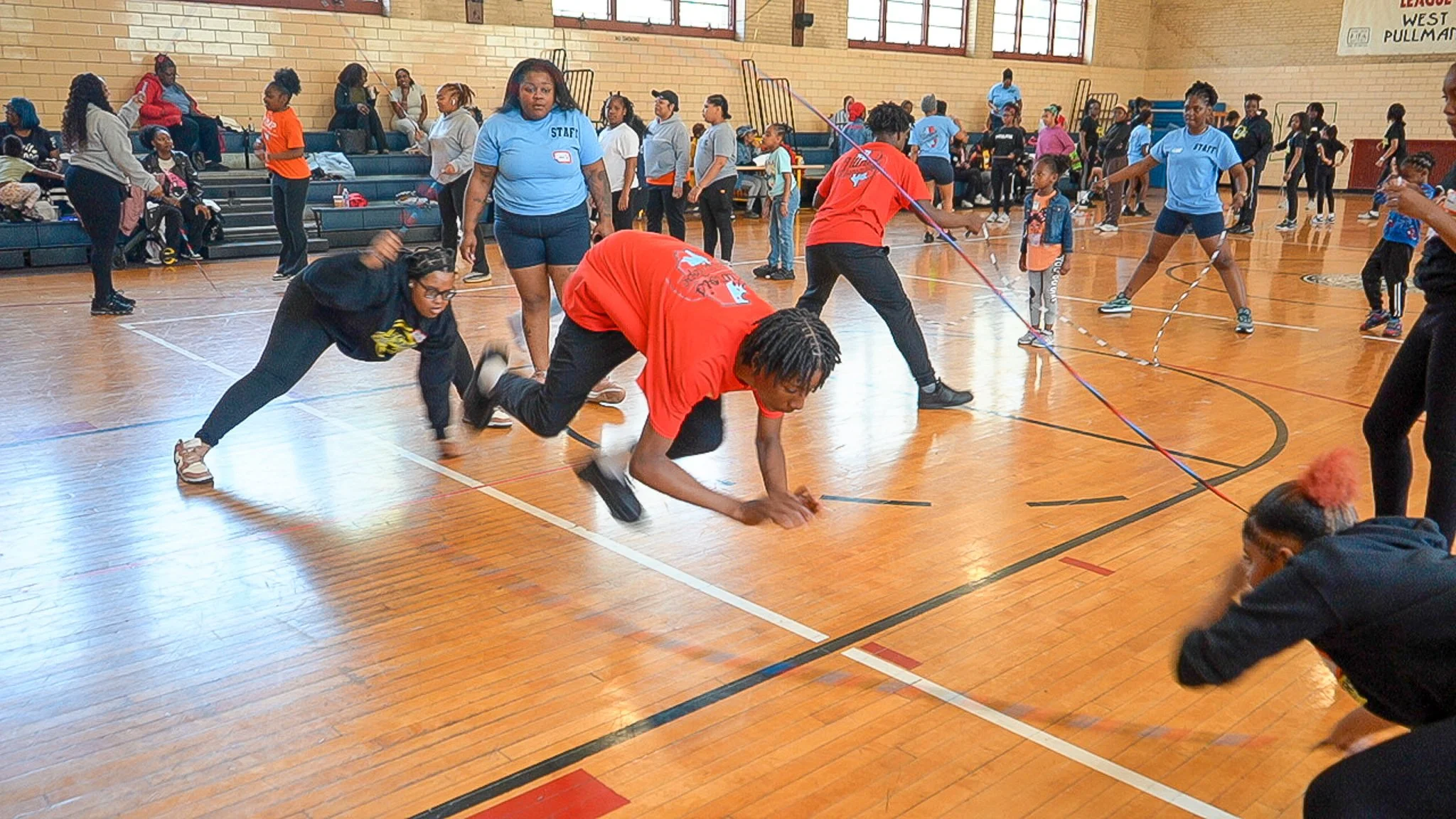 Group of children and adults in a gymnasium playing a game with jump ropes. Some children on the floor and some standing, with people sitting and standing on bleachers in the background.