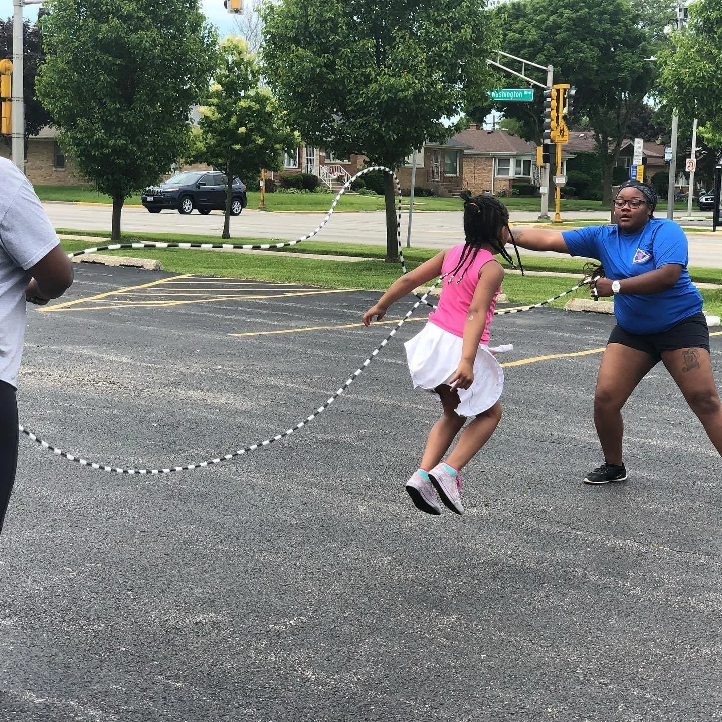 Coach LaTonya helping a young girl jump rope in a parking lot with trees, cars, and houses in the background.