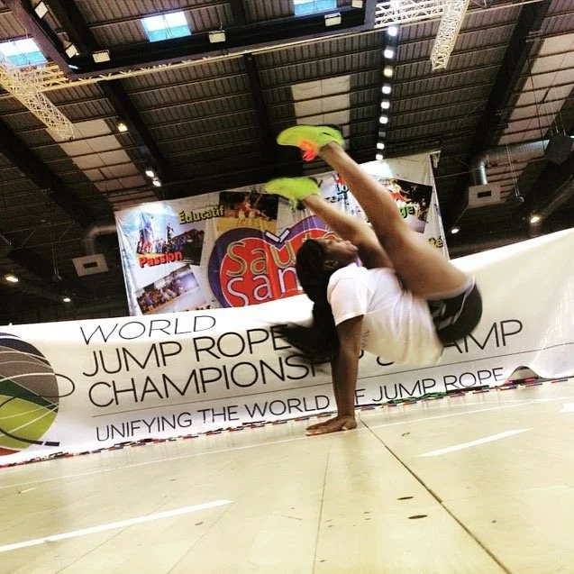 Female athlete performing a one-handed handstand with her legs bent back during a jump rope competition at an indoor sports arena.