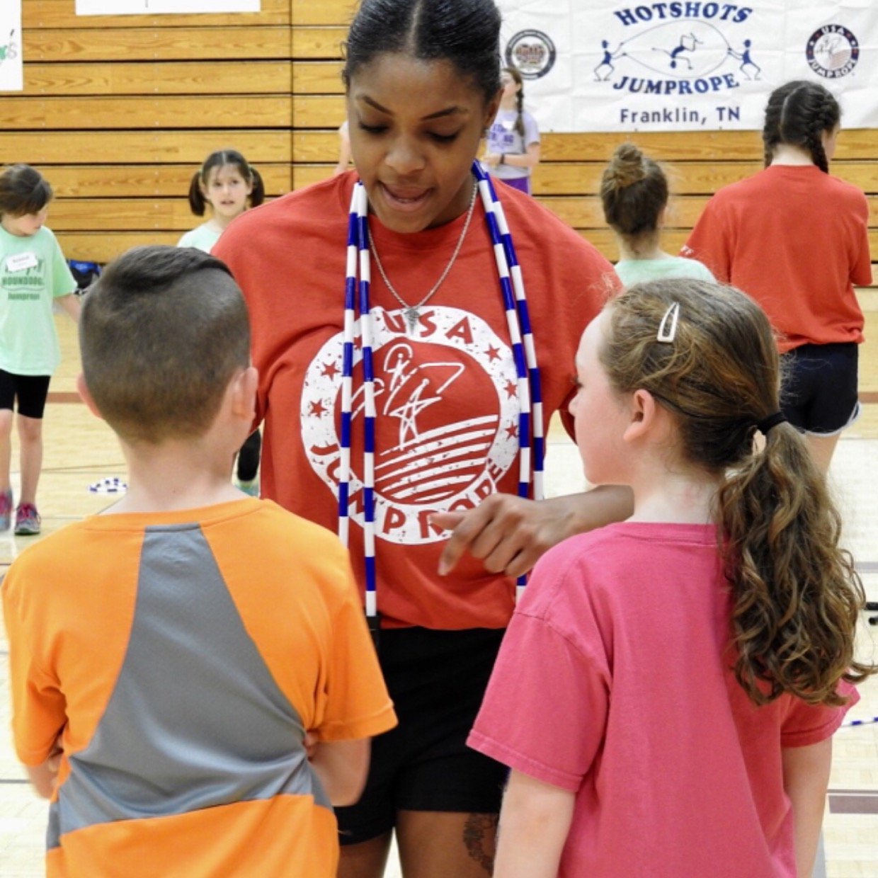 A woman wearing a red USA shirt and sports medals talks with two children, a boy and a girl, in a gym. There are other children and a banner in the background.