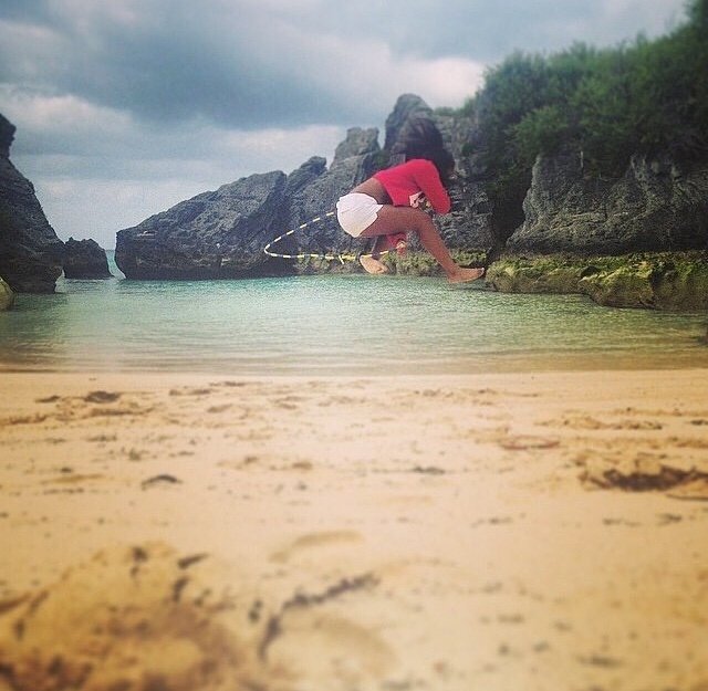 Person in a red shirt and white shorts crouching on rocks near the shoreline, with water surrounding the rocks and a cloudy sky overhead.