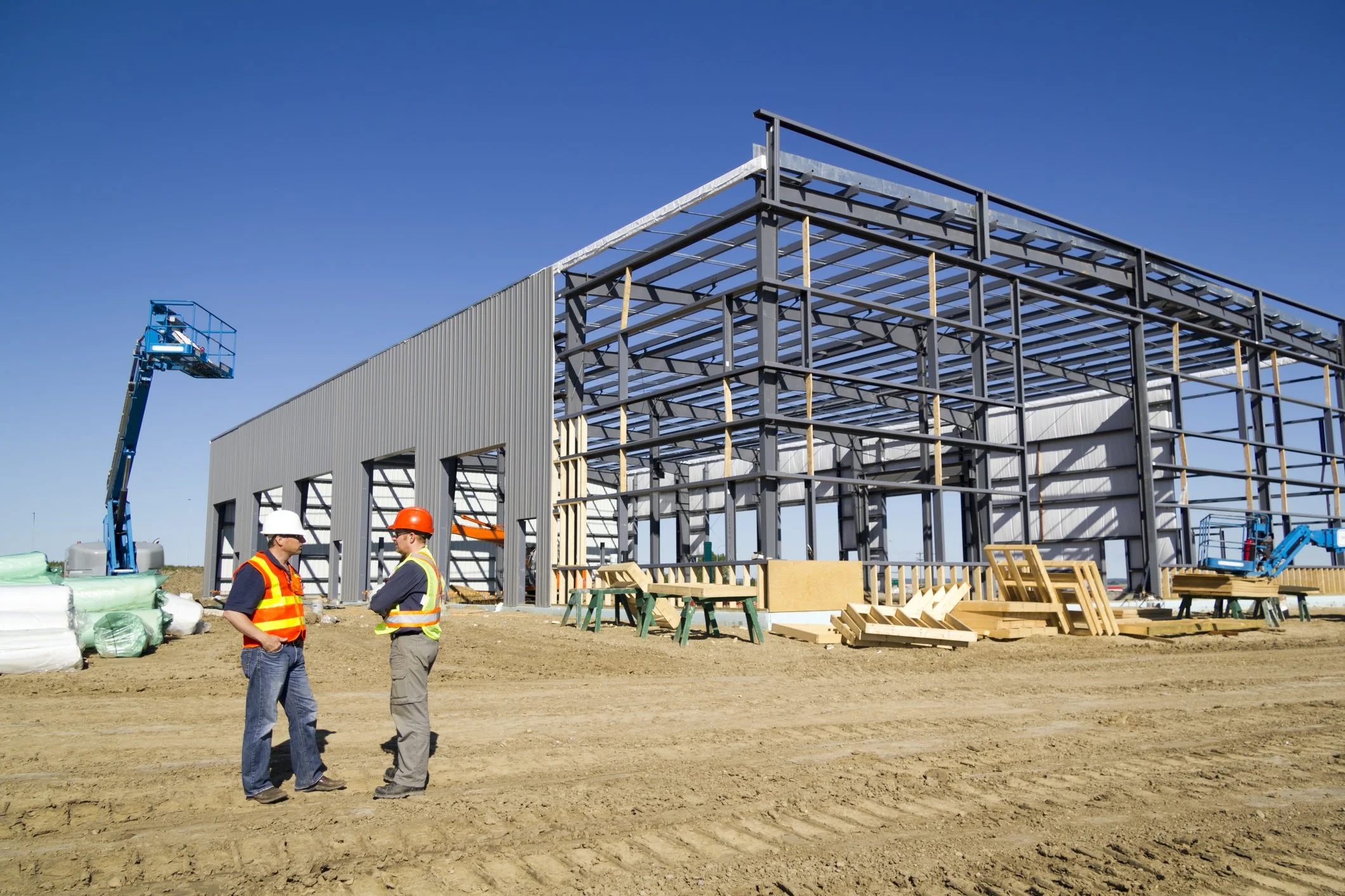 Construction site with steel framework for a building, two workers in safety vests and helmets talking, construction equipment and materials scattered around, clear blue sky.