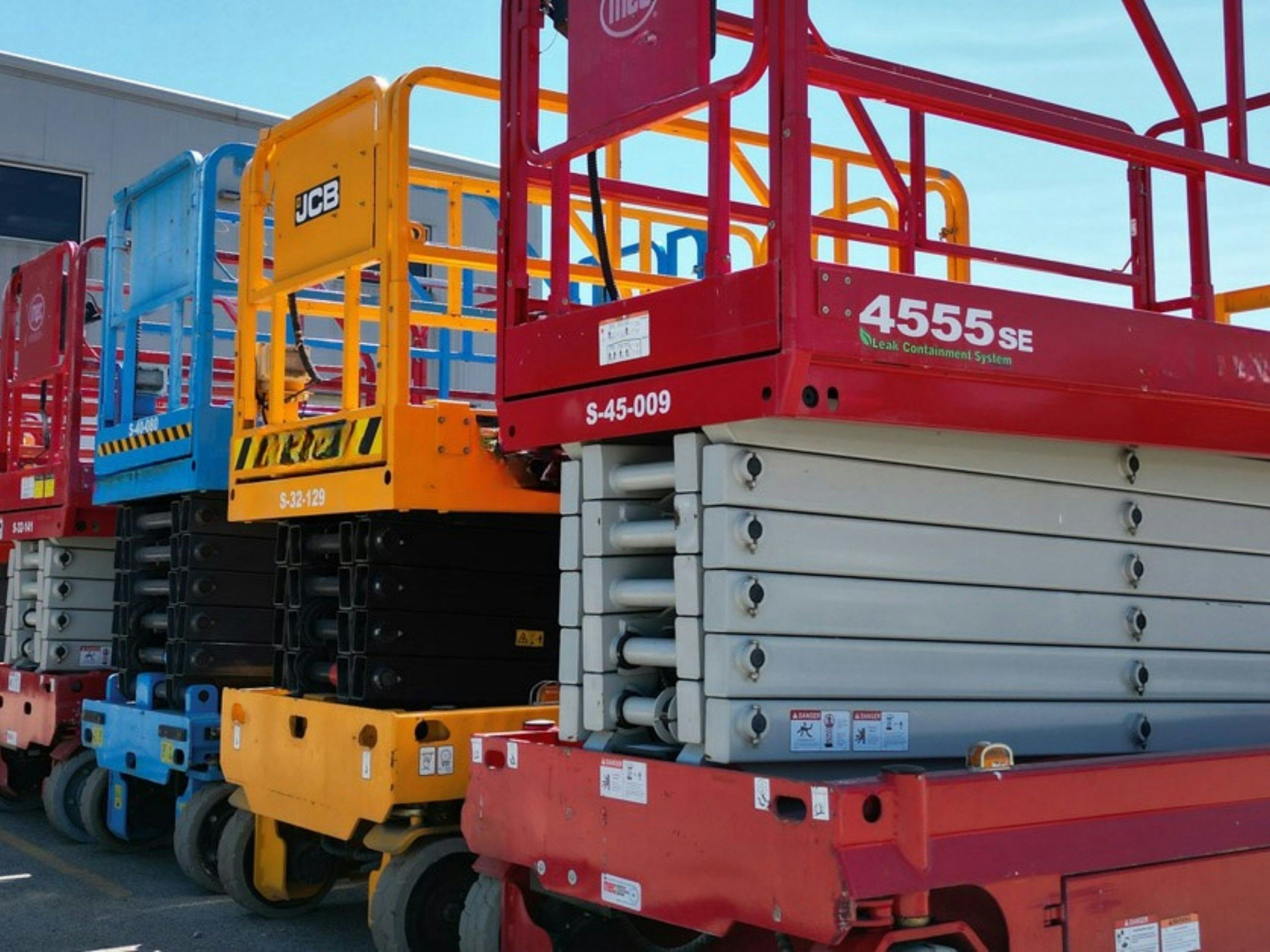 Stacked construction lifts or boom lifts in various colors including red, yellow, blue, and black, parked outdoors against a building with a clear sky.