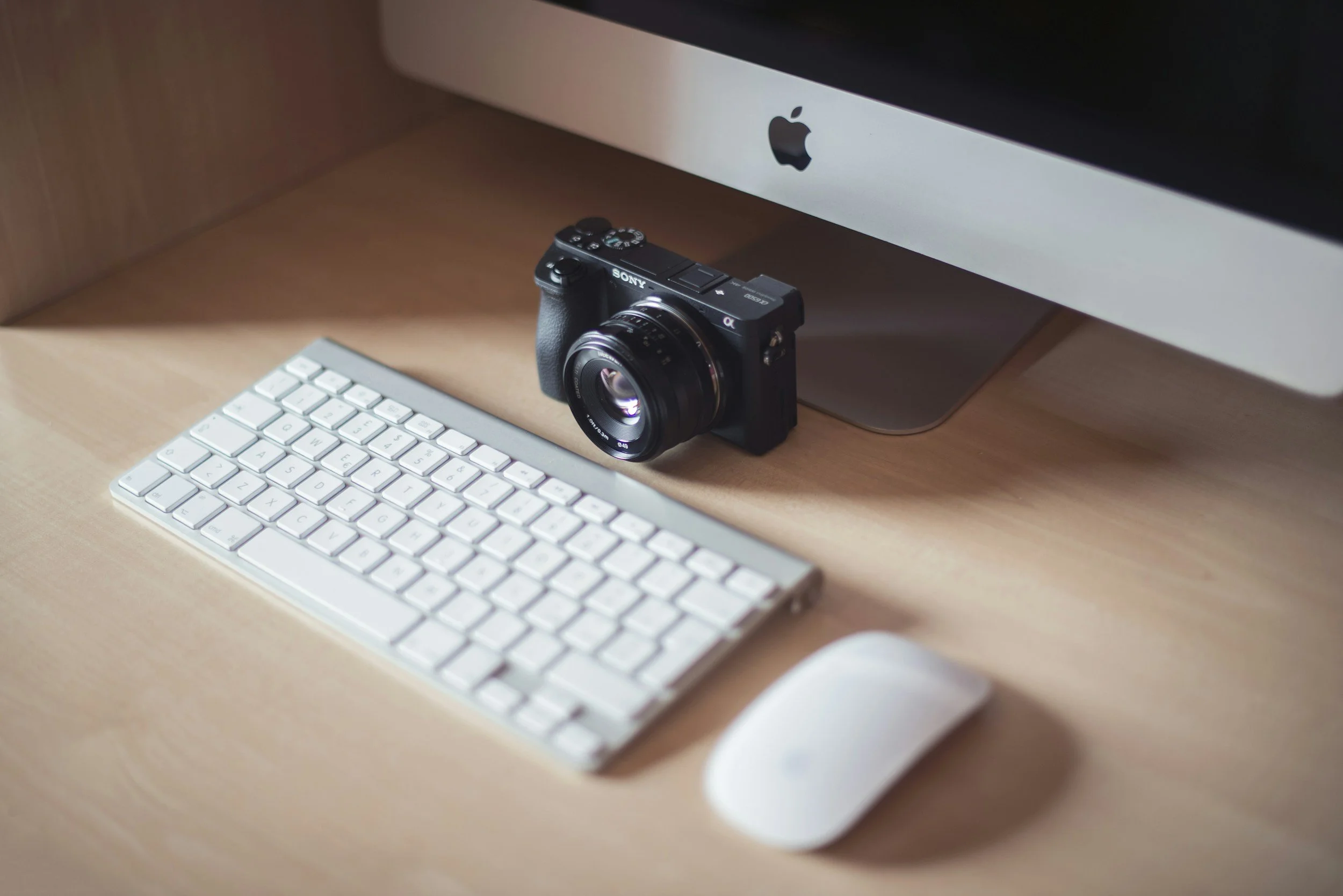 A high-angle view of a minimalist workspace featuring a Sony mirrorless camera, an Apple Magic Keyboard, and a Magic Mouse on a wooden desk beneath an iMac display.