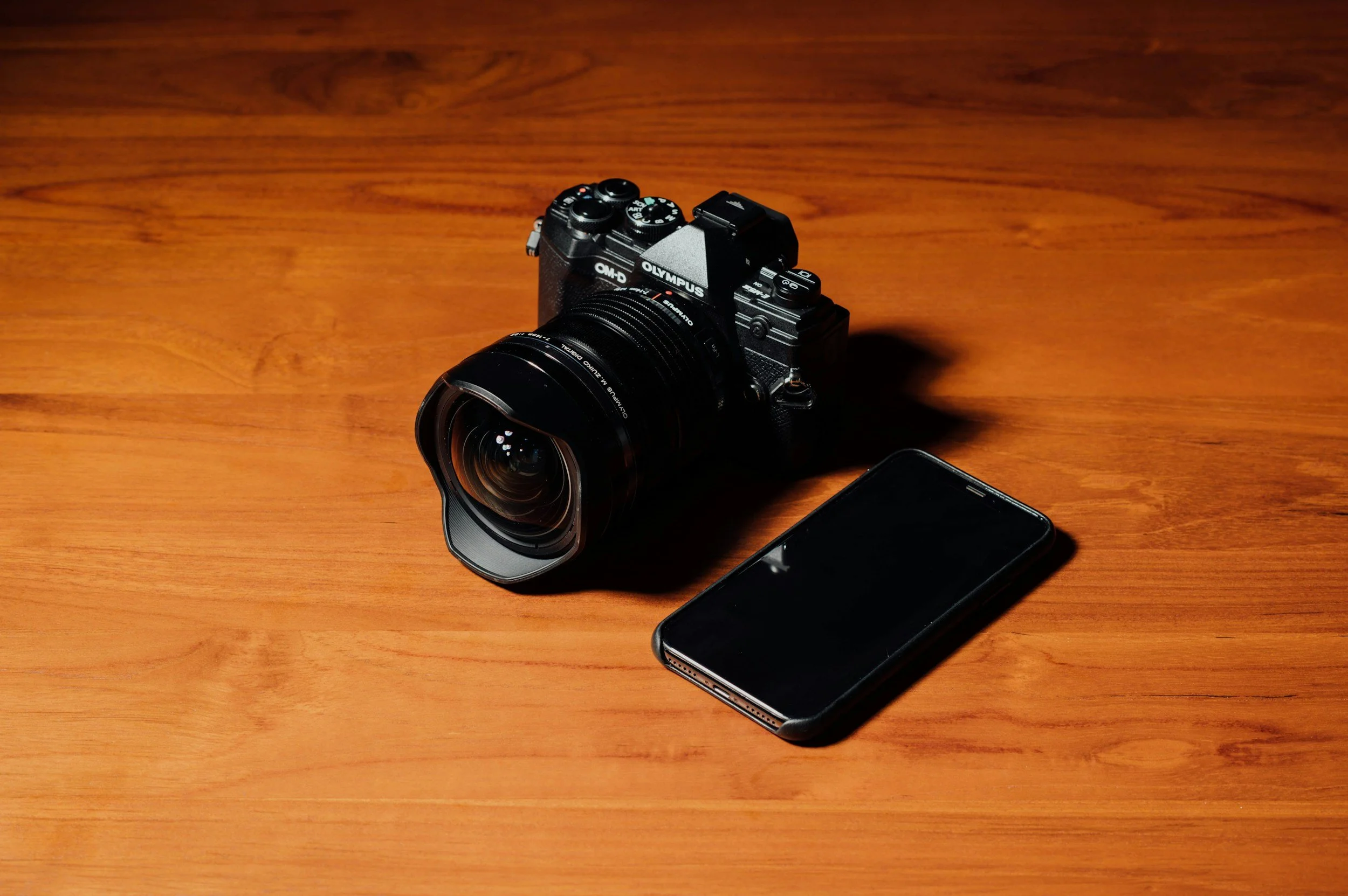 A high-angle studio shot of an Olympus OM-D mirrorless camera with a wide-angle lens sitting next to a black iPhone on a warm wood surface.