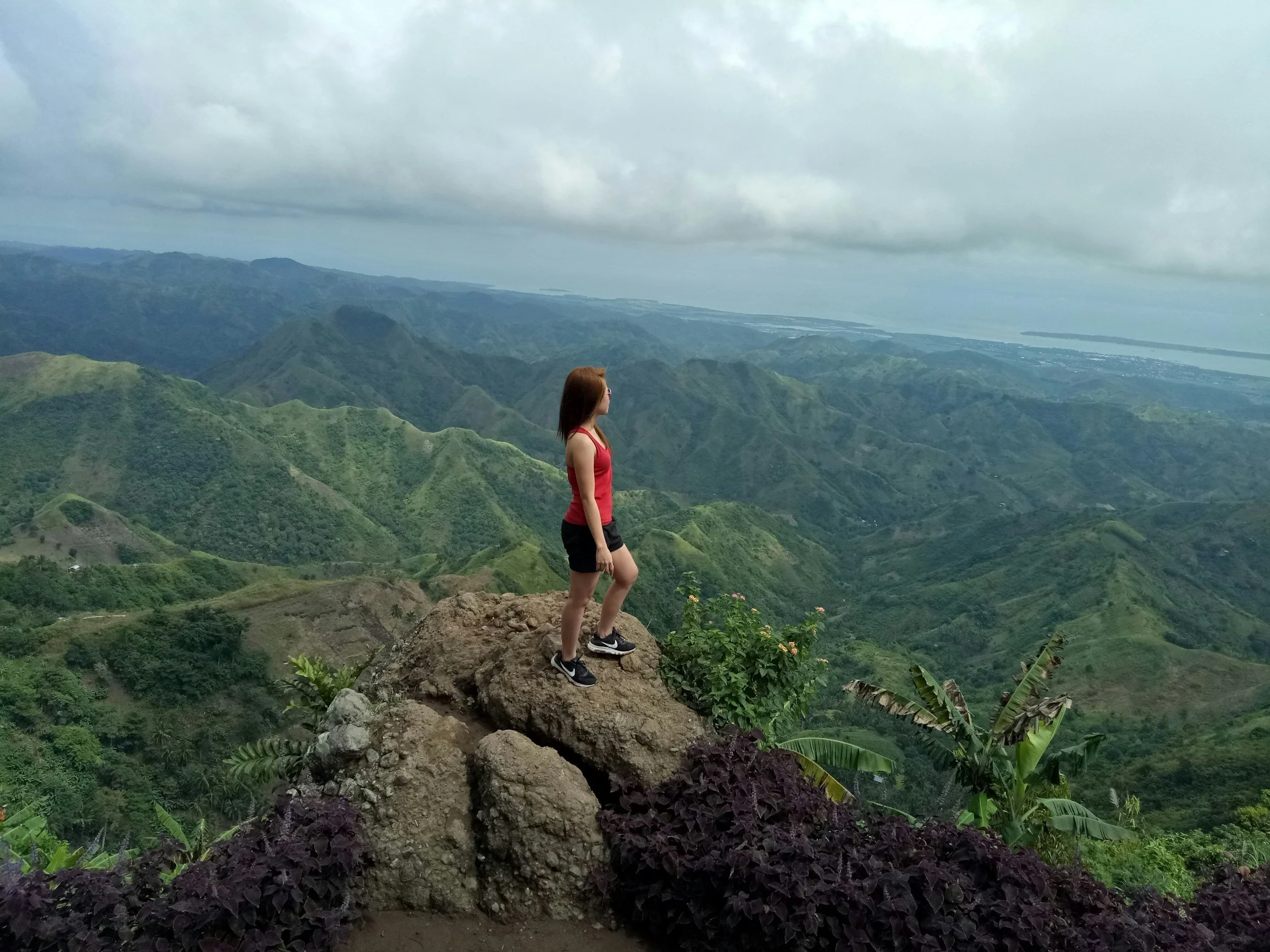 A woman with long hair, wearing a red tank top, black shorts, and sneakers, standing on a rocky outcrop overlooking lush green mountains and a cloudy sky.