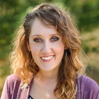 Close-up portrait of a young woman with curly blonde hair, blue eyes, smiling, outdoors with a blurred green background.