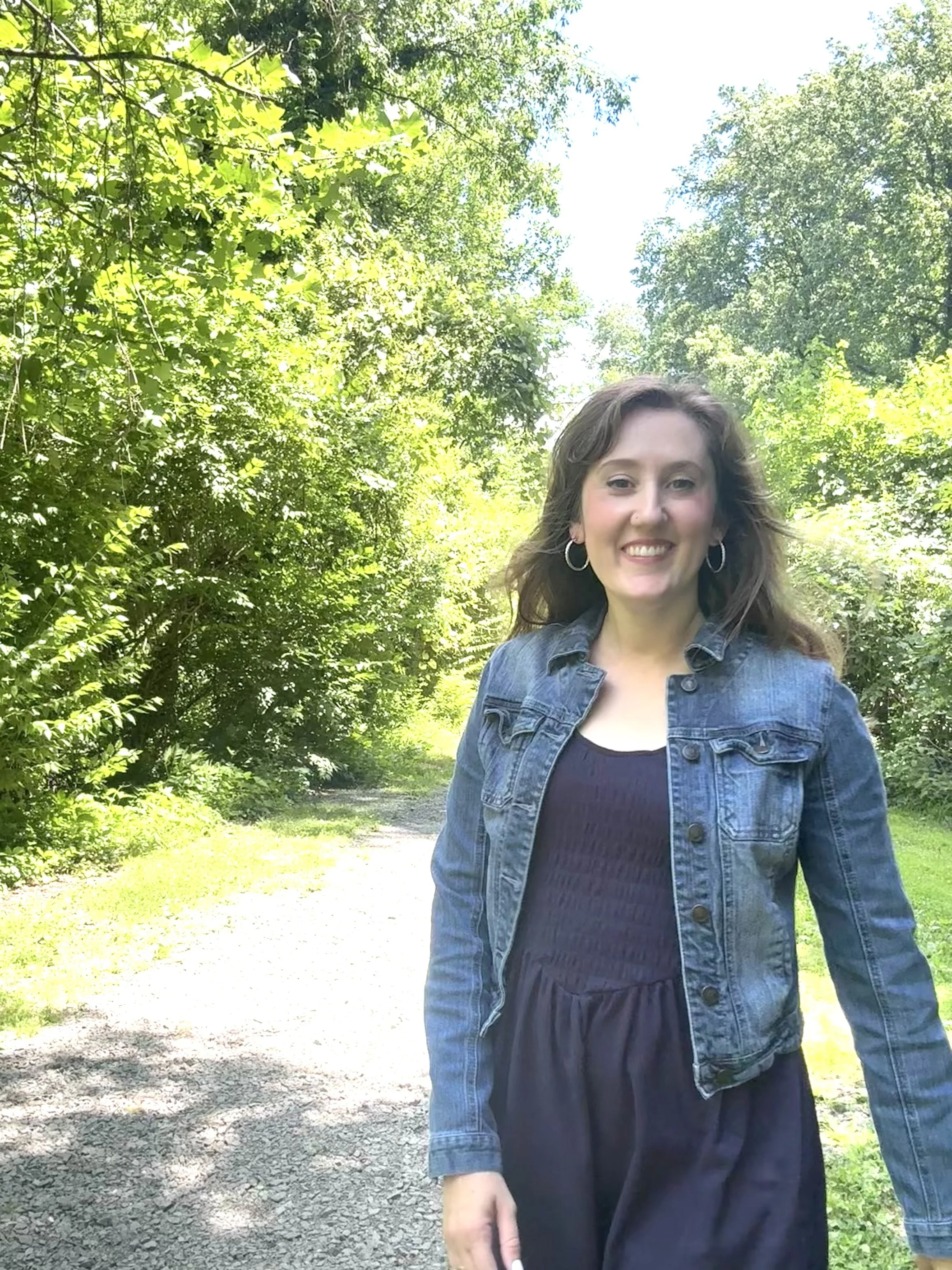 A woman walking outdoors on a tree-lined path, wearing a denim jacket over a dark dress, smiling at the camera.
