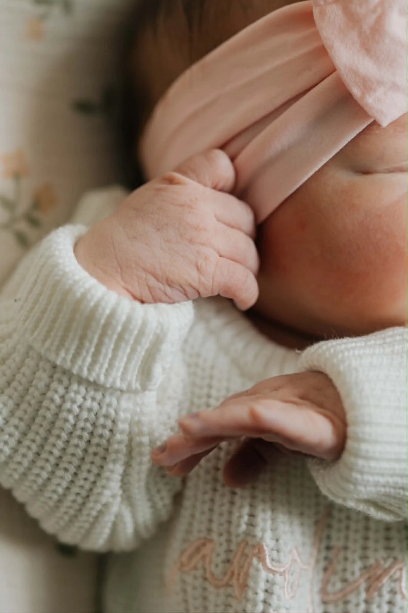 Close-up of a newborn baby with a pink headband, dressed in a white knitted sweater, covering their face with their hand.