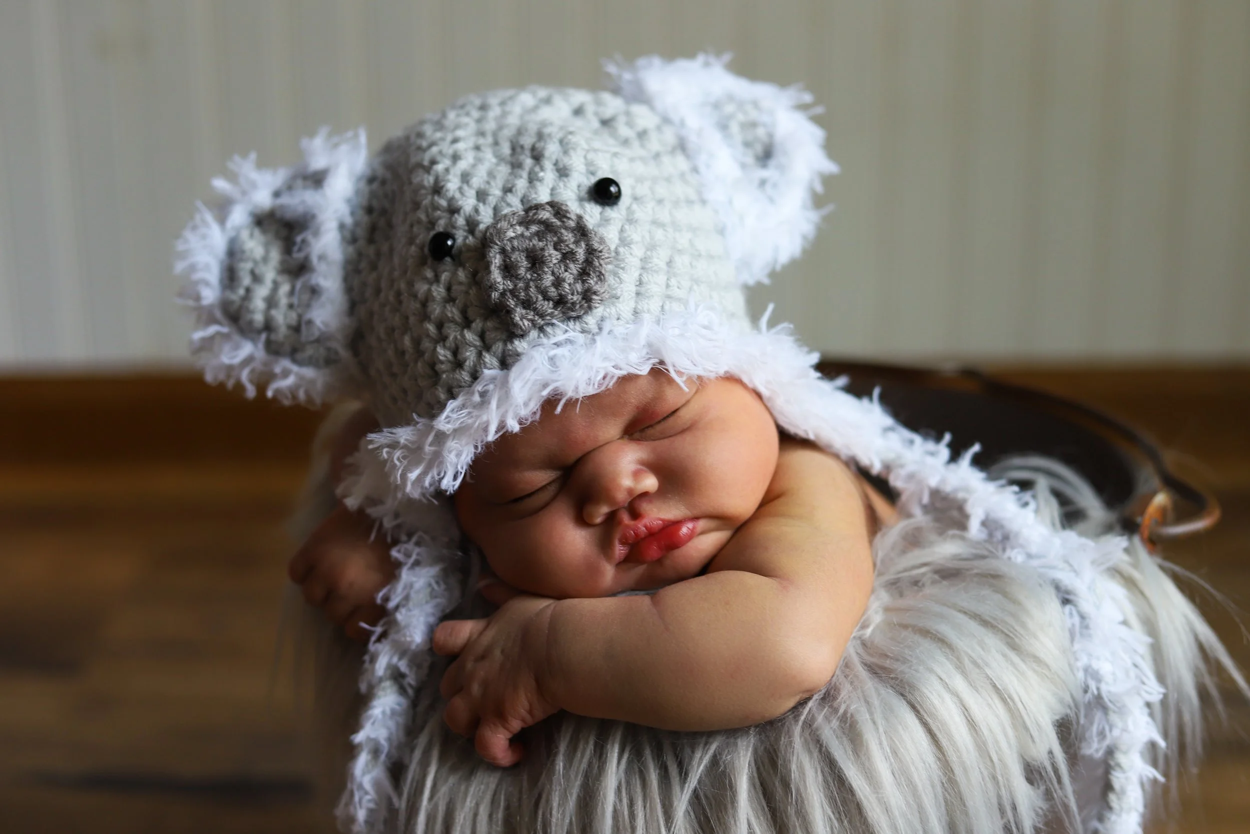 A sleeping newborn baby resting with arms crossed, wearing a cozy koala bear hat with floppy ears, black eyes, and a nose, on a fluffy blanket.