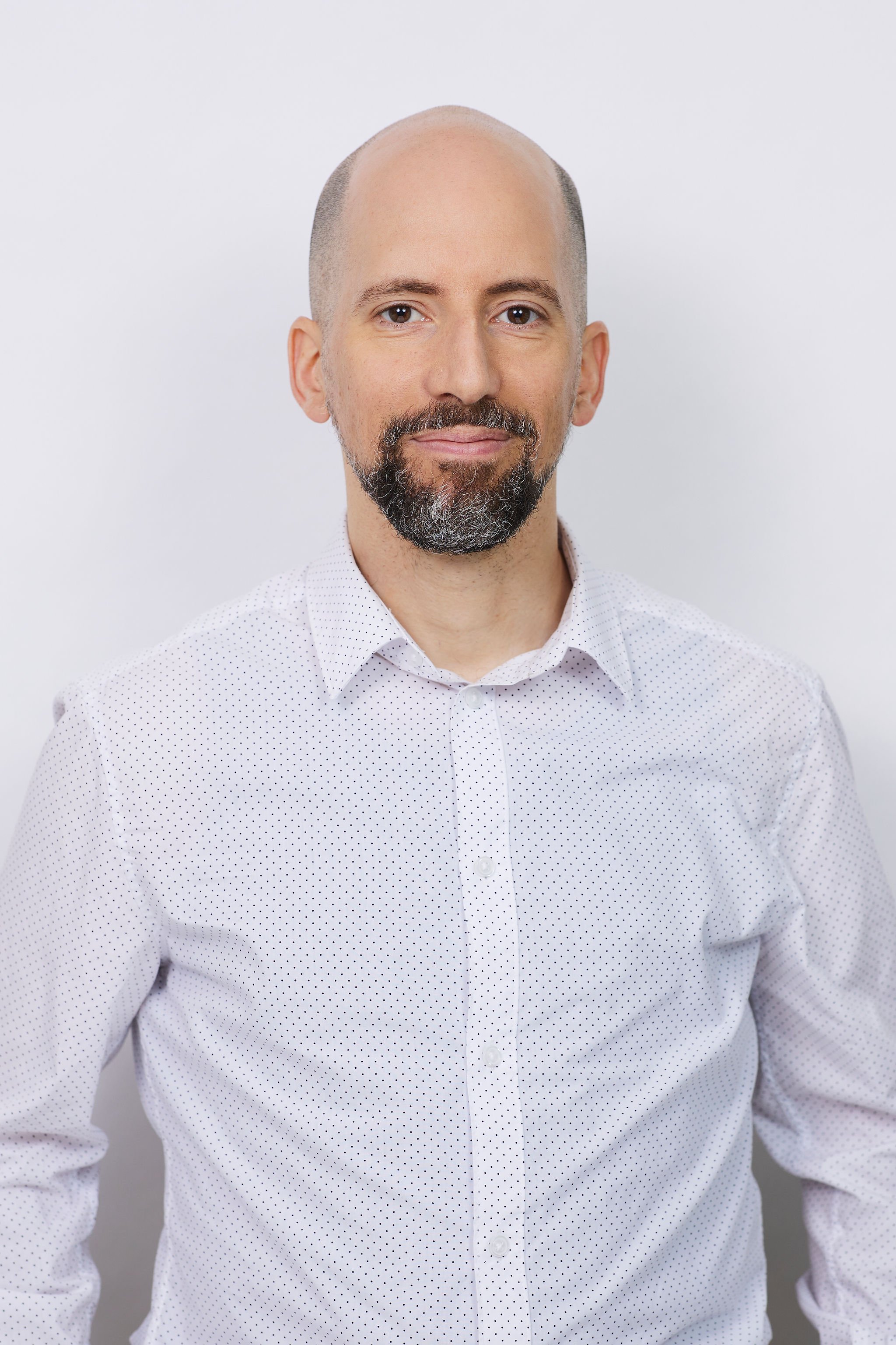 Portrait d'un homme avec une barbe, cheveux chauve, portant une chemise blanche à pois noirs, fond blanc.