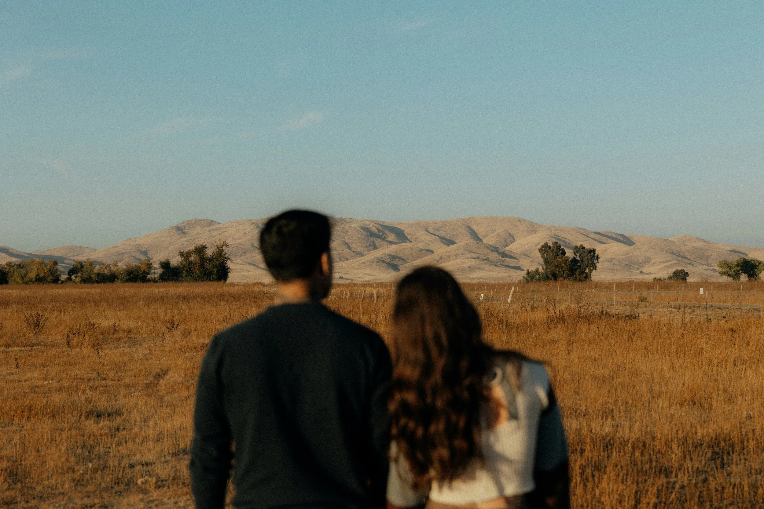 A group of people standing outdoors in a field, looking at mountains in the distance under a clear sky.