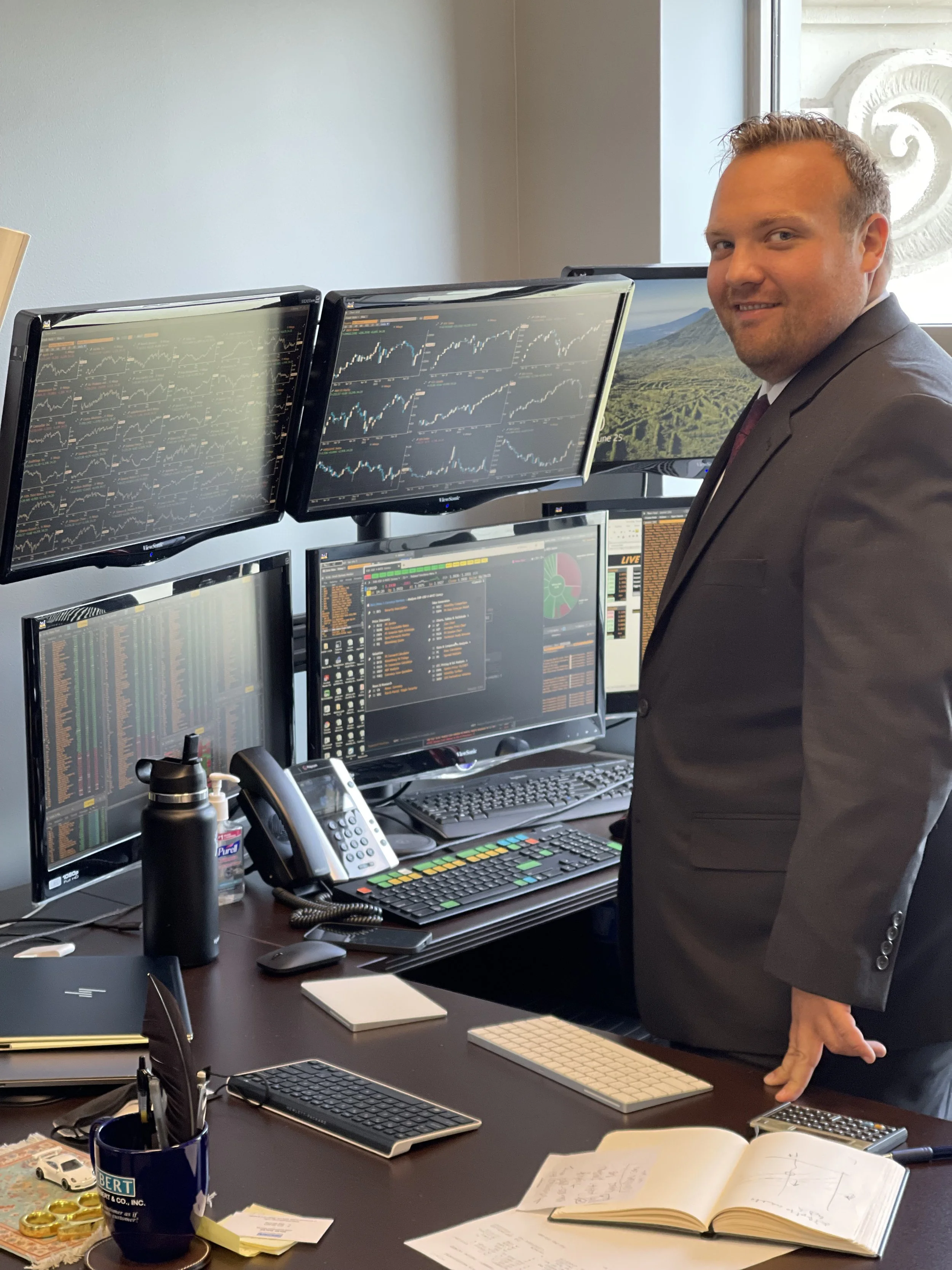John McGuinness in a suit standing in front of a Financial Professionals Desk with multiple computer monitors showing financial charts and data.