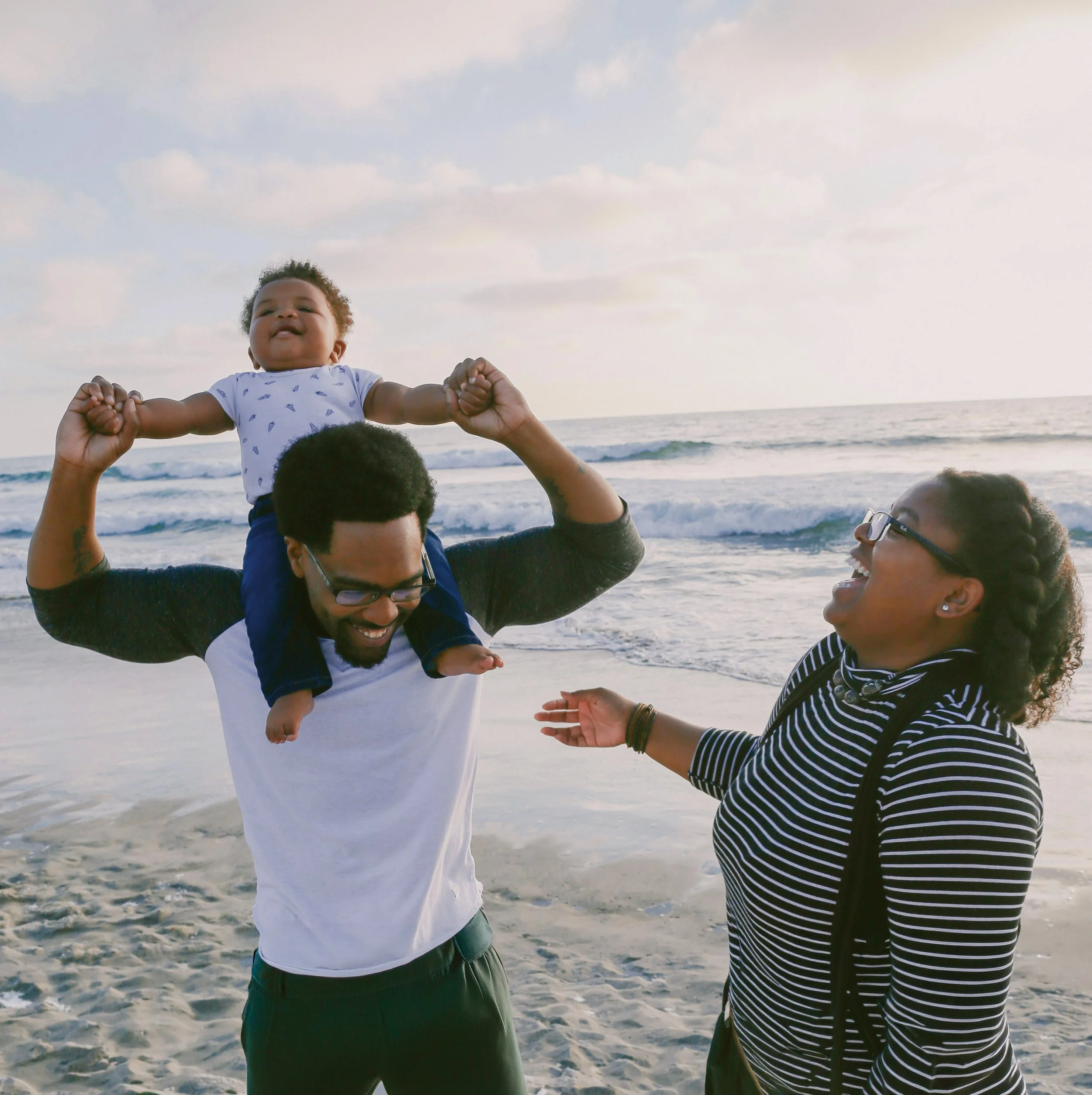 A group of people at the beach, with one child sitting on an adult's shoulders and others interacting, under a sky with clouds, ocean in the background.
