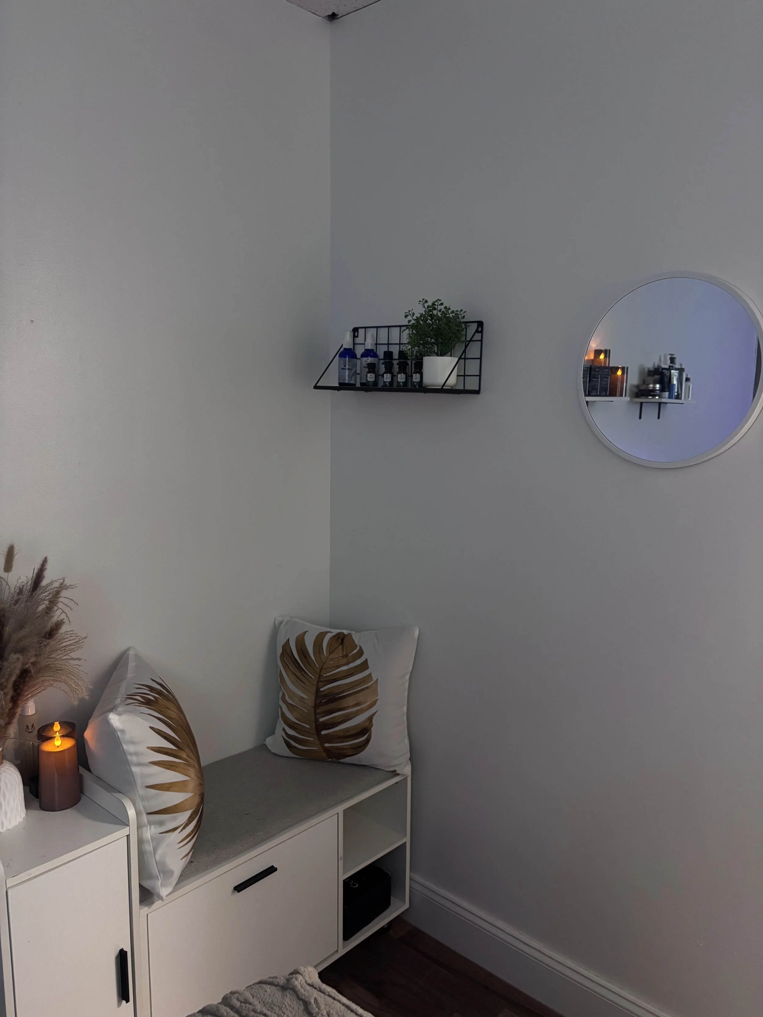 A corner of a room with a white cabinet, a decorative pillow with a leaf pattern, and candles. There is a wall shelf with bottles and a plant, and a round mirror reflecting other bottles and candles.