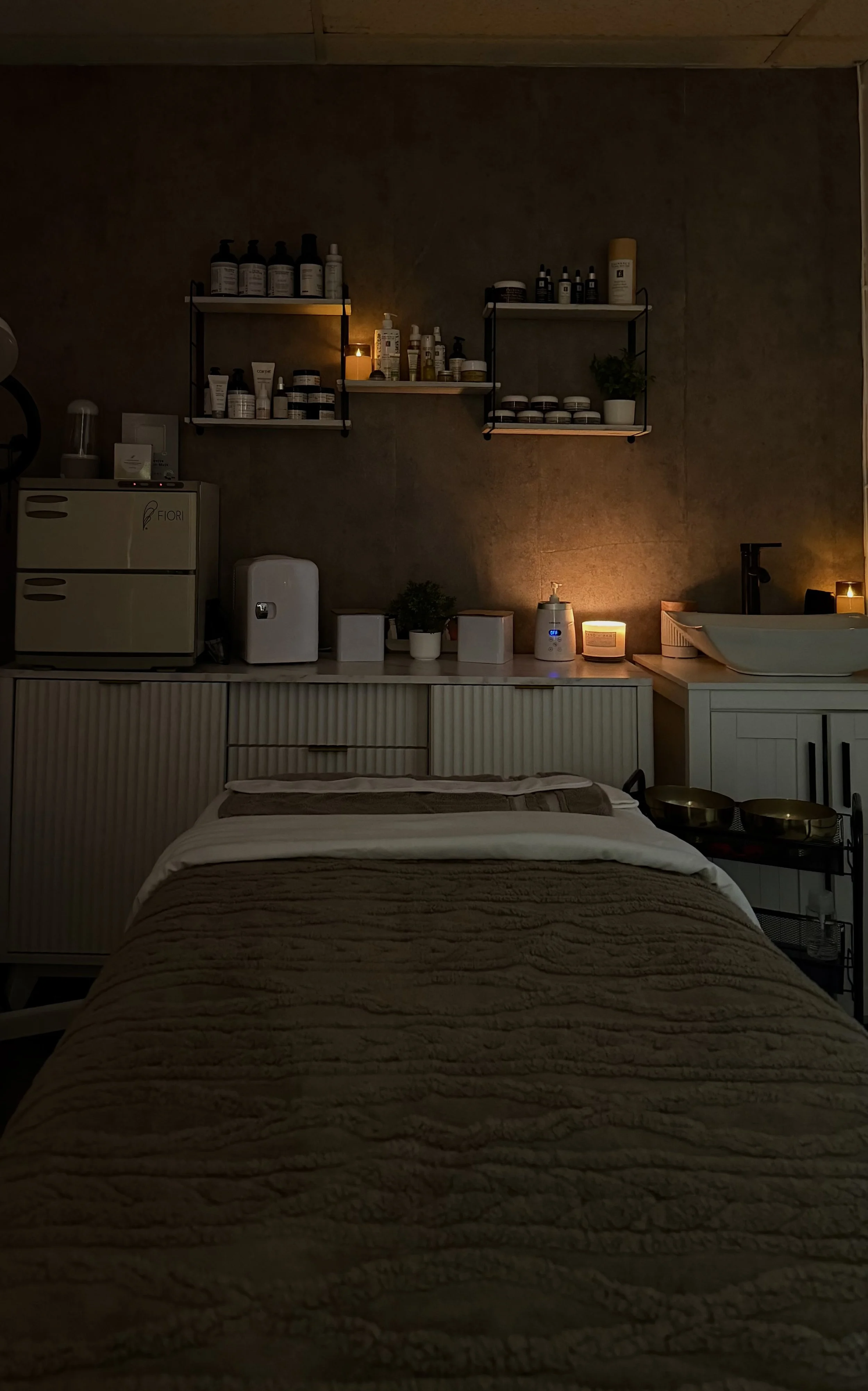 A cozy, dimly lit wellness or treatment room with a bed in the foreground, covered with a beige textured blanket. The background features shelves with skincare or beauty products, a small sink, and a counter with various items including a humidifier and small potted plants. Soft lighting creates a relaxing atmosphere.