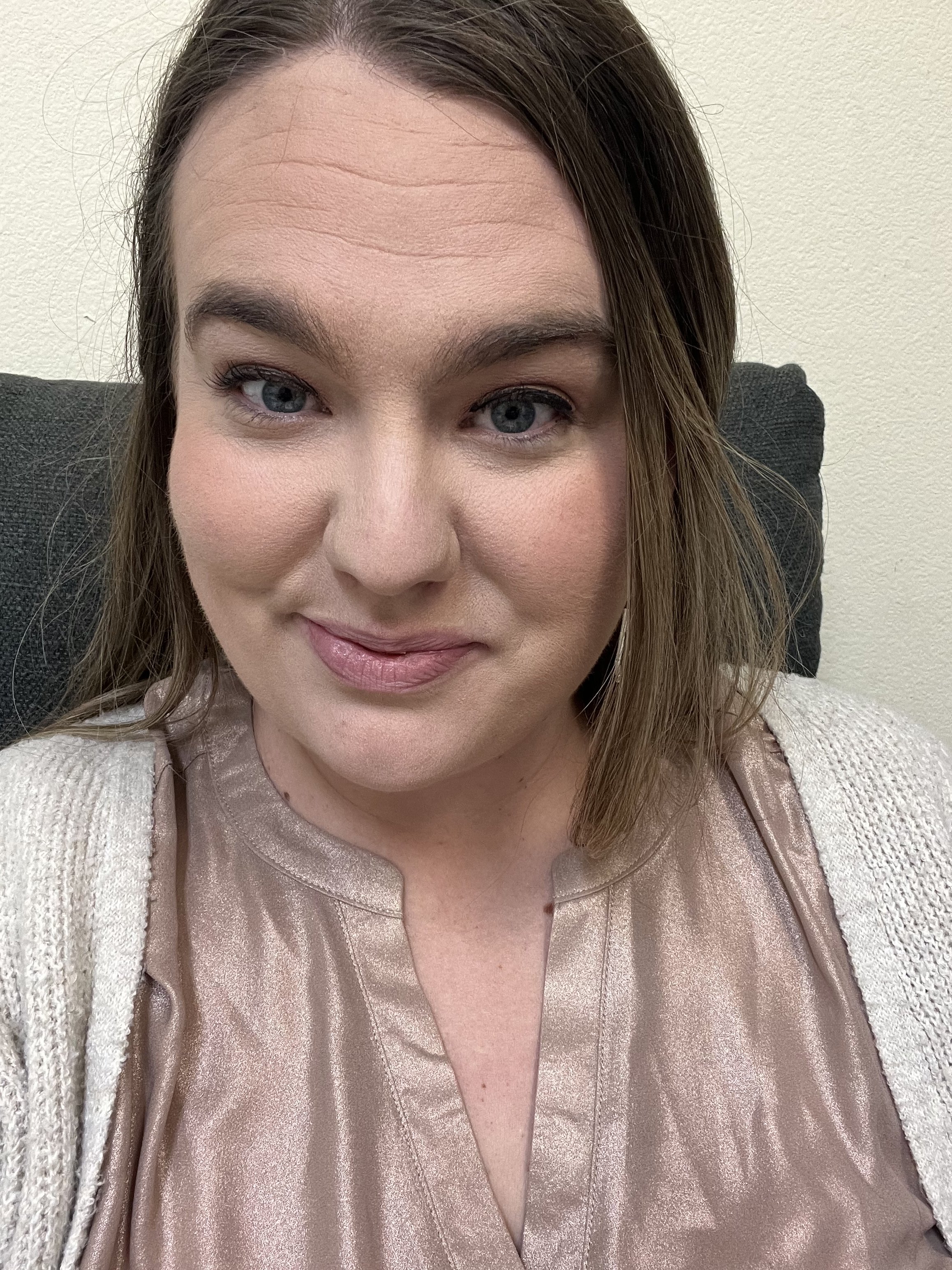 A woman with light skin and brown hair partially tied back, smiling subtly, wearing a shiny, peach-colored satin blouse with a V-neck and a cream-colored knitted cardigan.