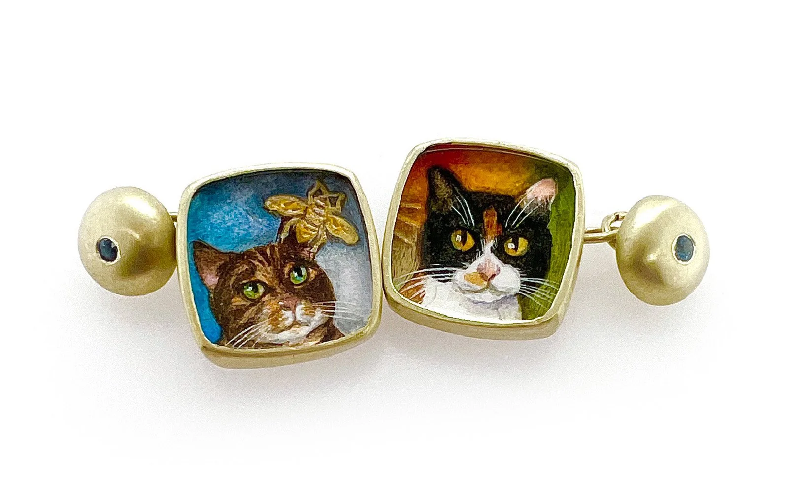 Cufflinks with two square frames featuring painted portraits of cats, one with a bee on its head, and two gold beads with small blue gems.