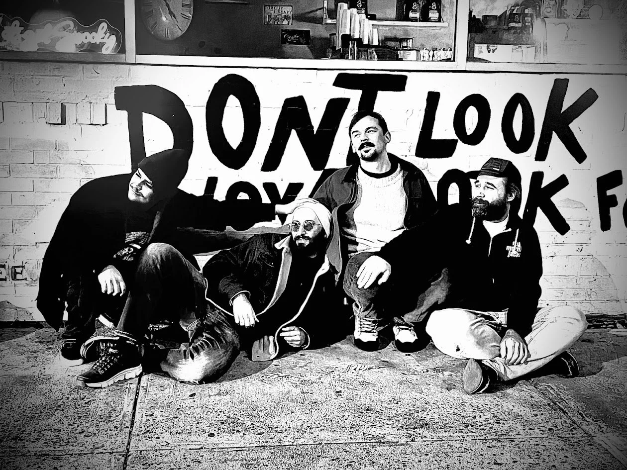 Four men sit and lie on the ground in front of a wall with graffiti that says 'DON'T LOOK' and other words partially visible. They are dressed casually and appear to be friends posing for a photo.
