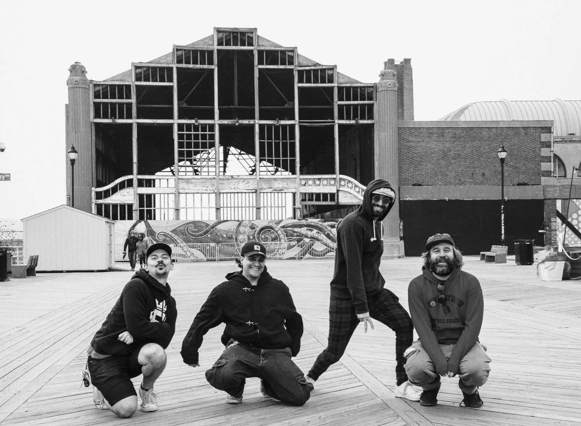 A group of five smiling men posing on a wooden boardwalk in front of a partially constructed building with a large, rounded roof. Some of the men are squatting while others stand, all dressed in casual clothes and hoodies. There are street lamps and a colorful painted wall behind them.