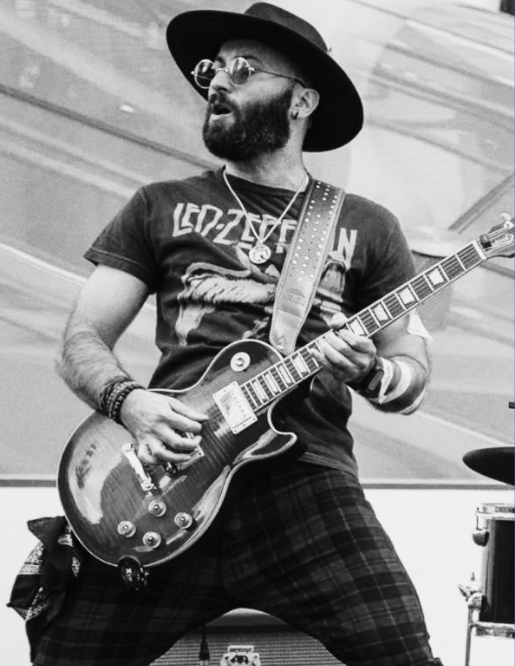 Musician with a beard, wearing glasses, a wide-brimmed hat, a Led Zeppelin T-shirt, and plaid pants, playing an electric guitar on stage.