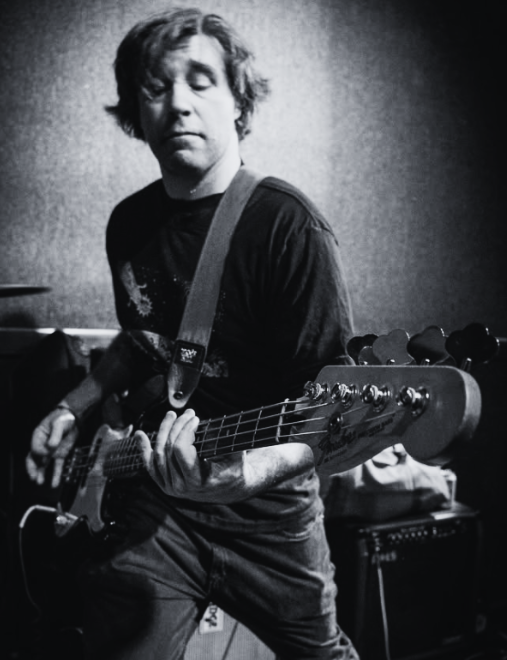 A man with shoulder-length wavy hair playing an electric bass guitar in a music studio, with a drum set partially visible in the background.