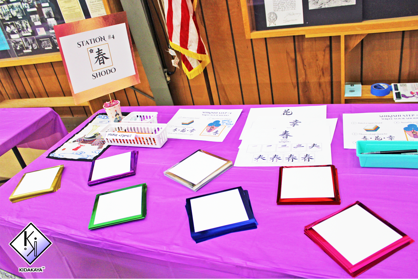 A table with a pink tablecloth displays colorful stacks of paper, a small white basket with practice pencils, and instruction sheets for practicing Japanese characters. A sign on a stick reads 'STATION #4' and 'SHODO' with a Japanese character. An Am