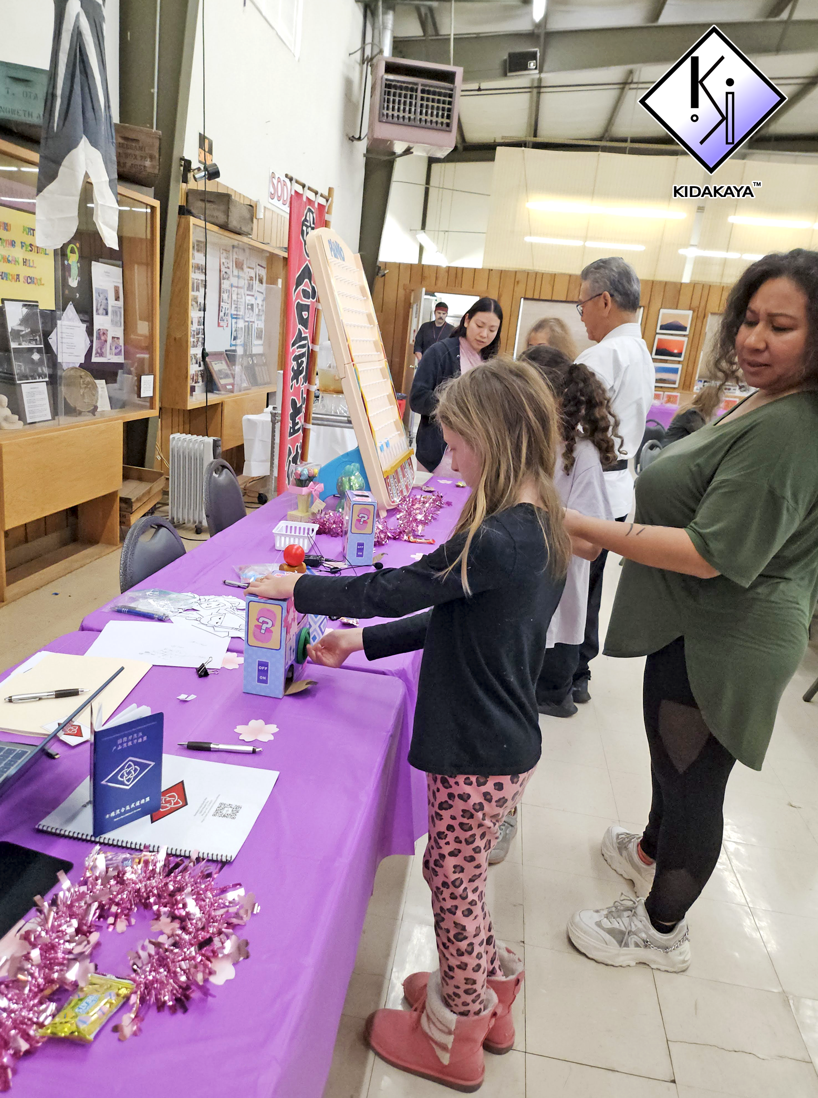 A group of children and adults participating in an event at a table covered with a purple cloth. The table has various small items, decorations, and papers. The background shows wooden panels and informational posters. One girl wearing pink leopard-p